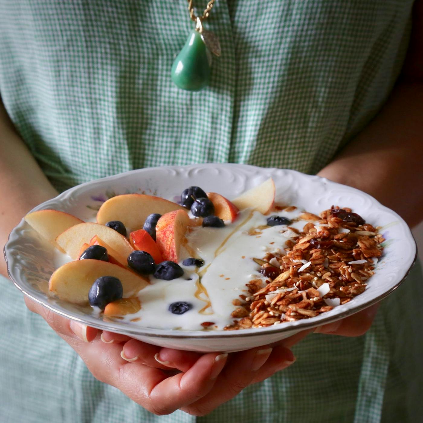 Person holding bowl of granola with tahini and silan along with fresh fruit and yogurt.