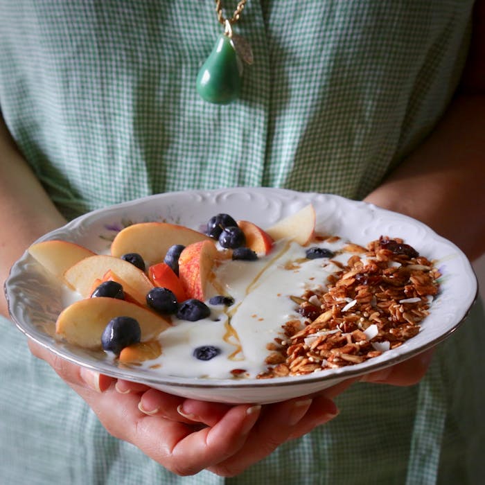 Person holding bowl of granola with tahini and silan along with fresh fruit and yogurt.