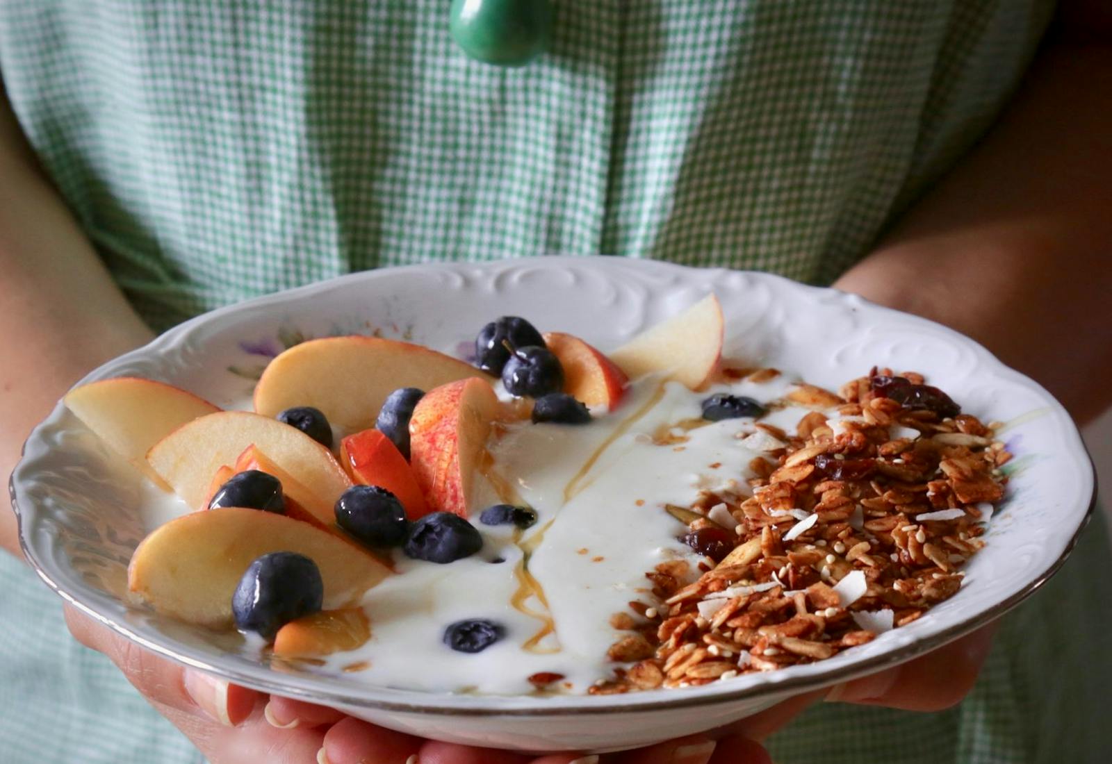 Person holding bowl of granola with tahini and silan along with fresh fruit and yogurt.