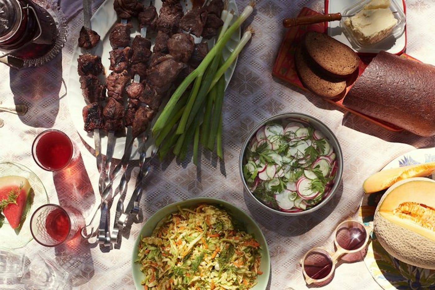 Lamb shish kebobs surrounded by red wine, melon, rye bread and salads.