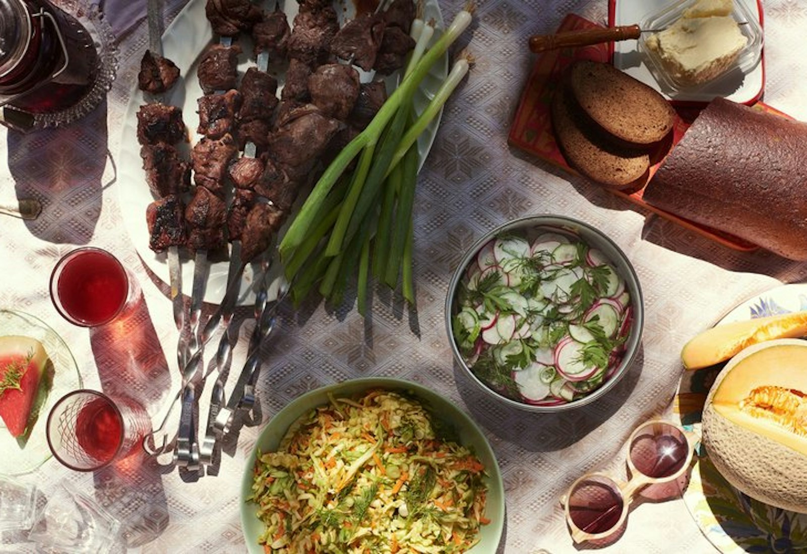 Lamb shish kebobs surrounded by red wine, melon, rye bread and salads.