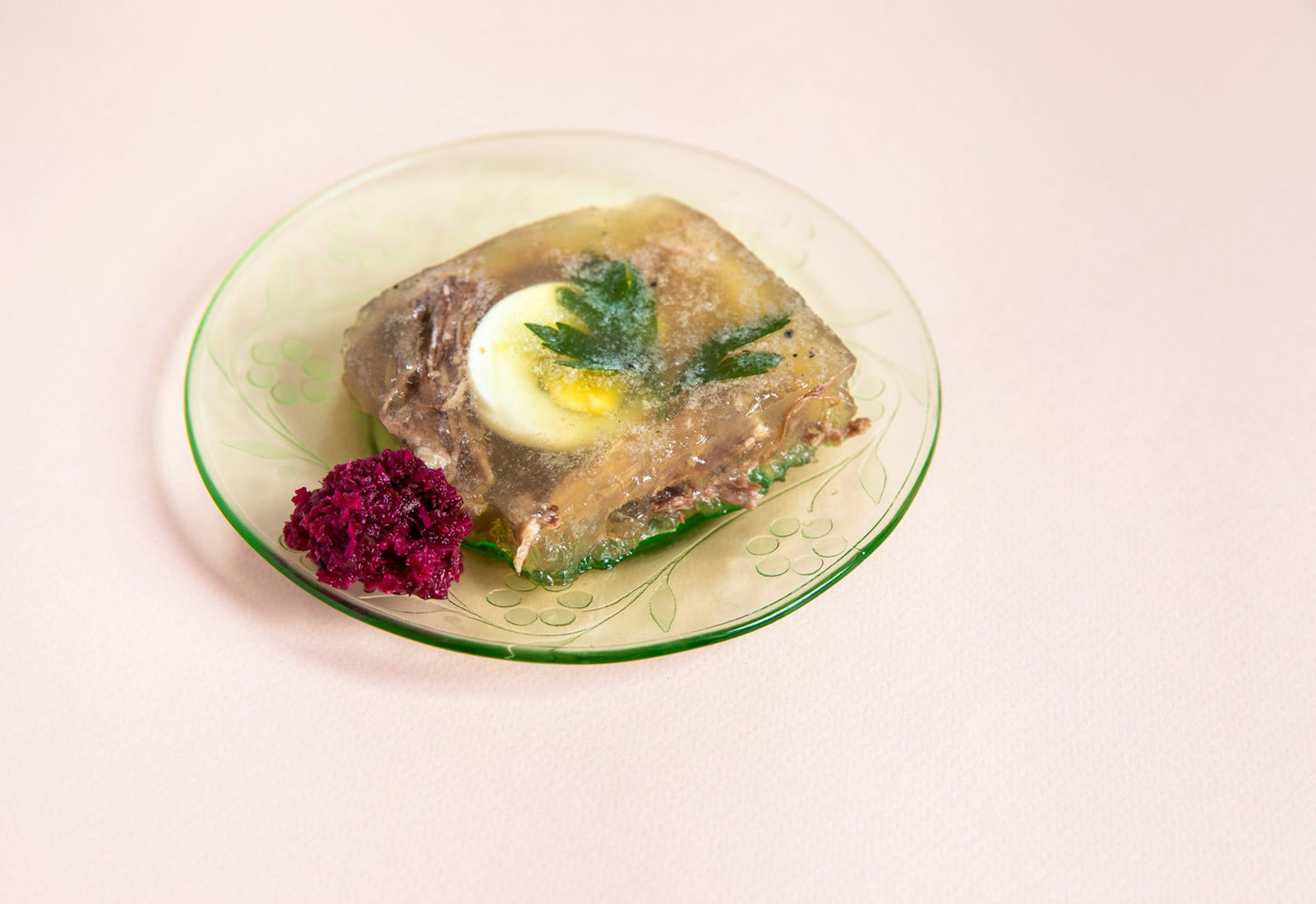 Slice of p'tcha with dollop of beet horseradish on glass plate atop pale pink surface.