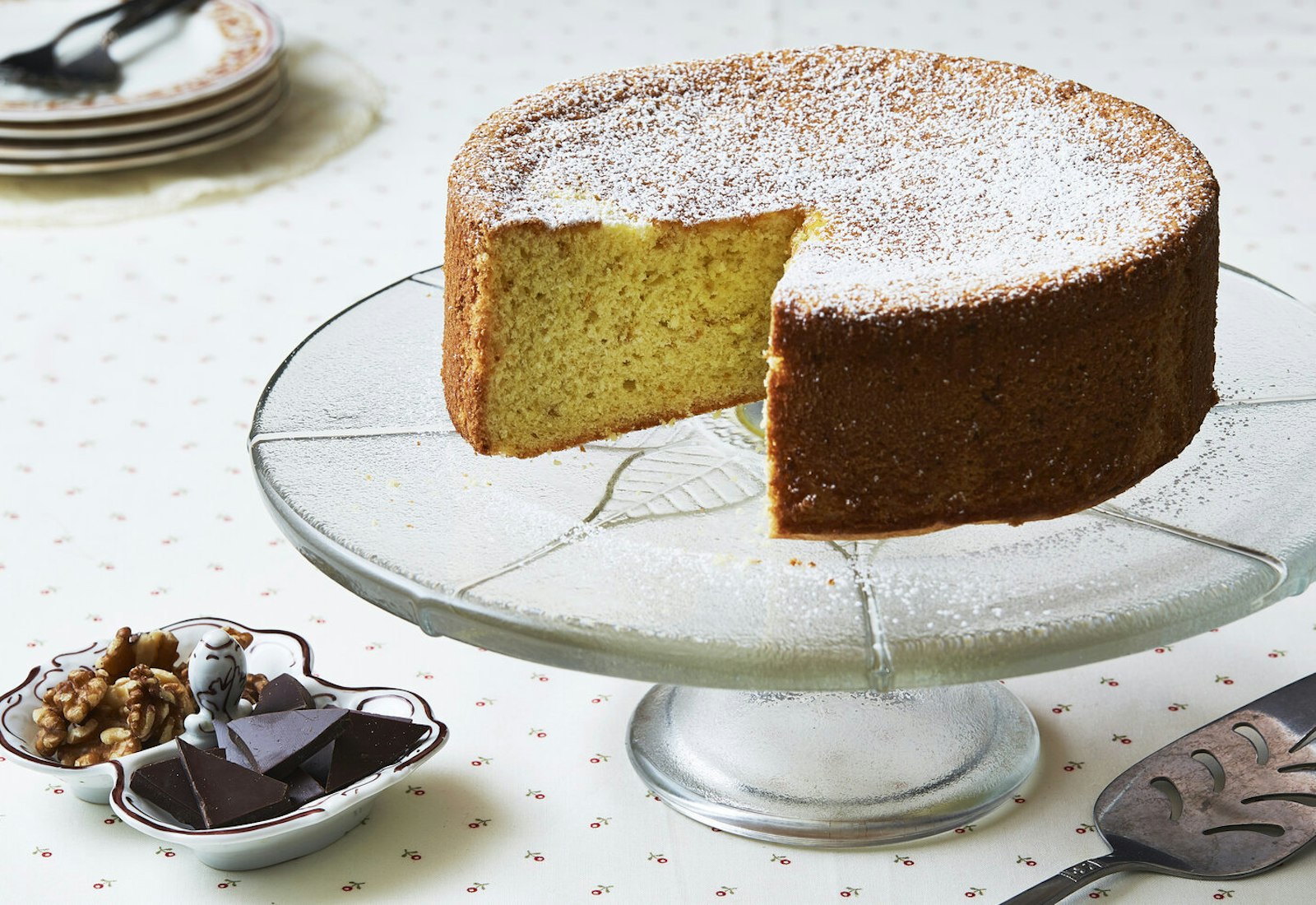 Biscuit on cake tray with slice removed, dish with walnuts and chocolate.