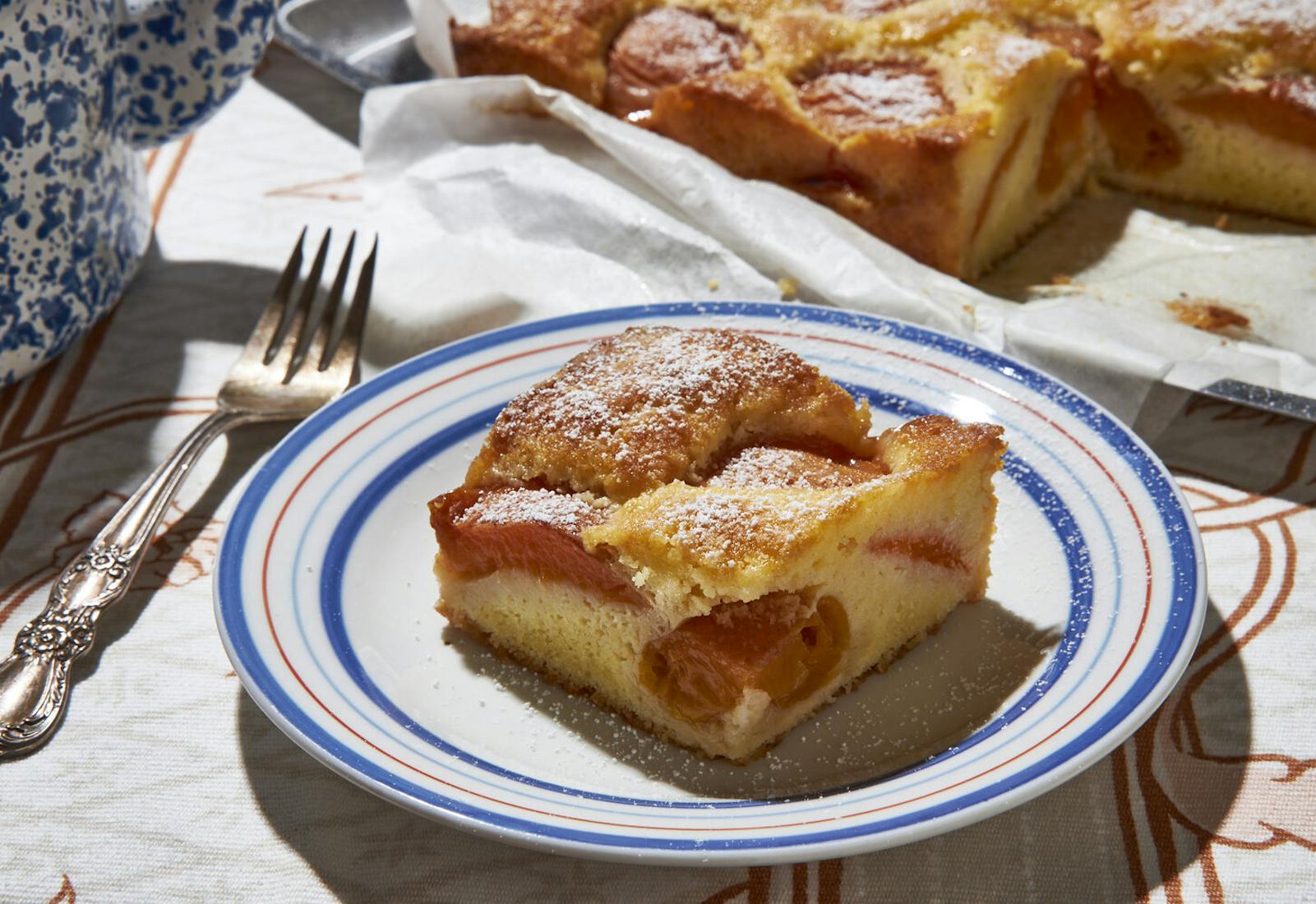 Slice of apricot cake on red and blue striped plate alongside whole cake on pan.