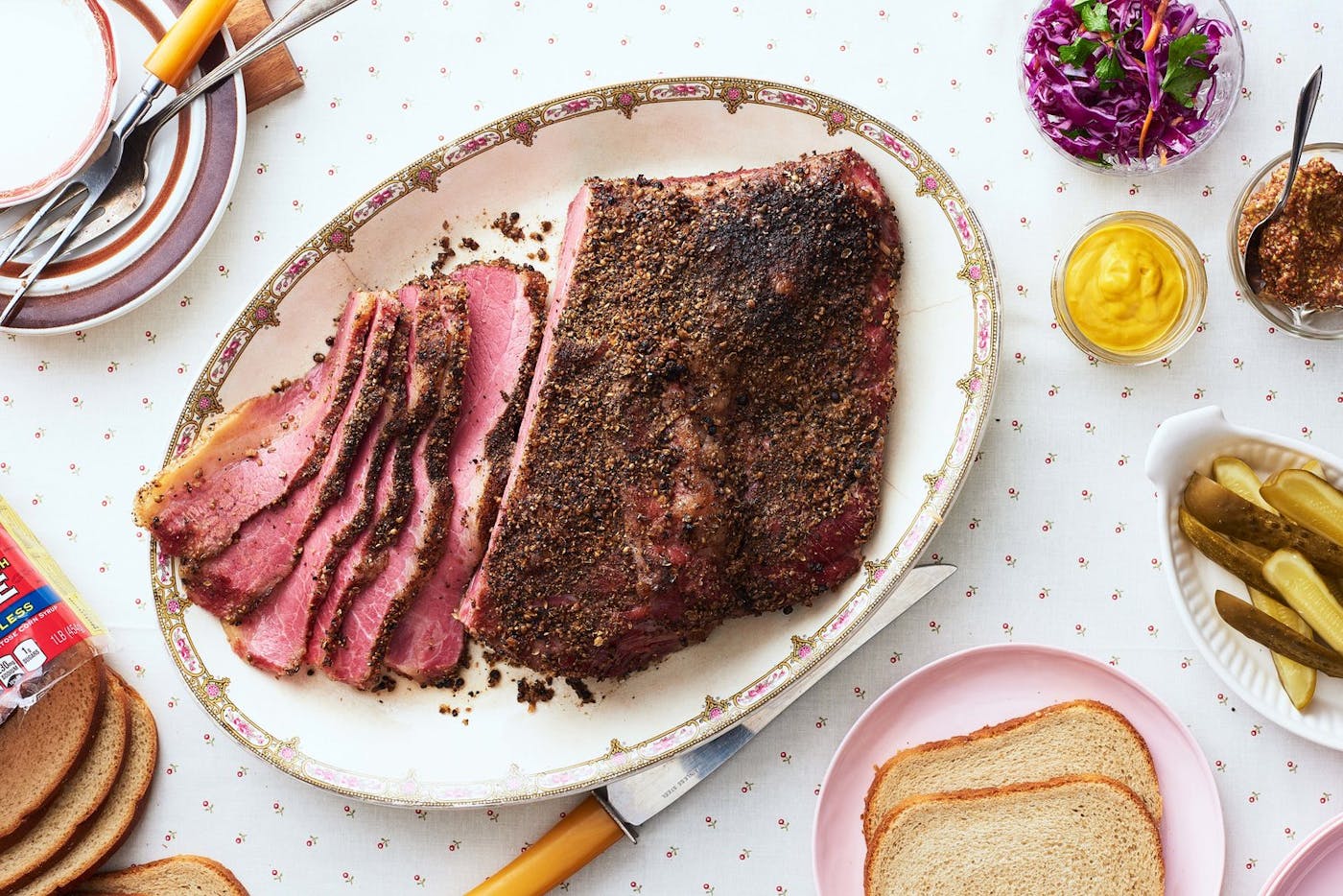 Partially sliced pastrami on oblong plate alongside grainy and smooth mustard, pickles, sliced bread, and purple slaw atop flower-dotted tablecloth.