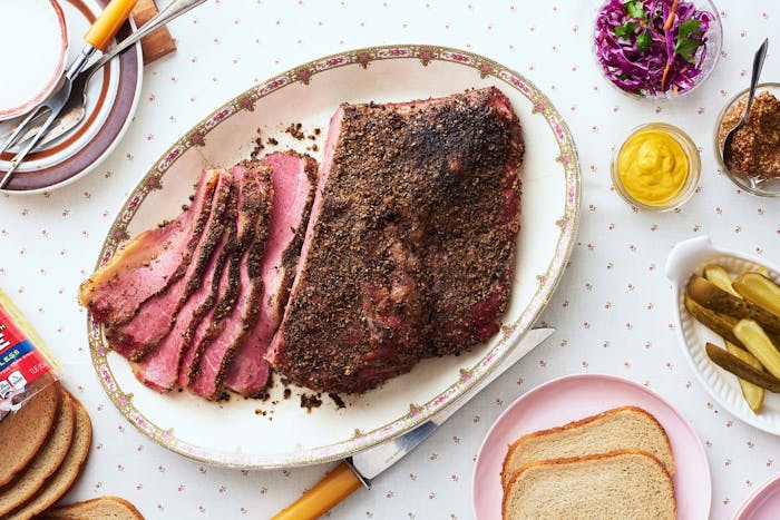 Partially sliced pastrami on oblong plate alongside grainy and smooth mustard, pickles, sliced bread, and purple slaw atop flower-dotted tablecloth.