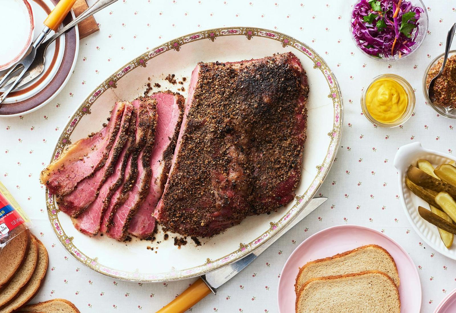 Partially sliced pastrami on oblong plate alongside grainy and smooth mustard, pickles, sliced bread, and purple slaw atop flower-dotted tablecloth.