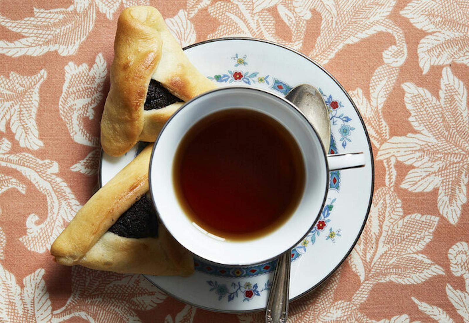 Poppy seed hamantaschen on plate with cup of tea, atop dark pink tablecloth.