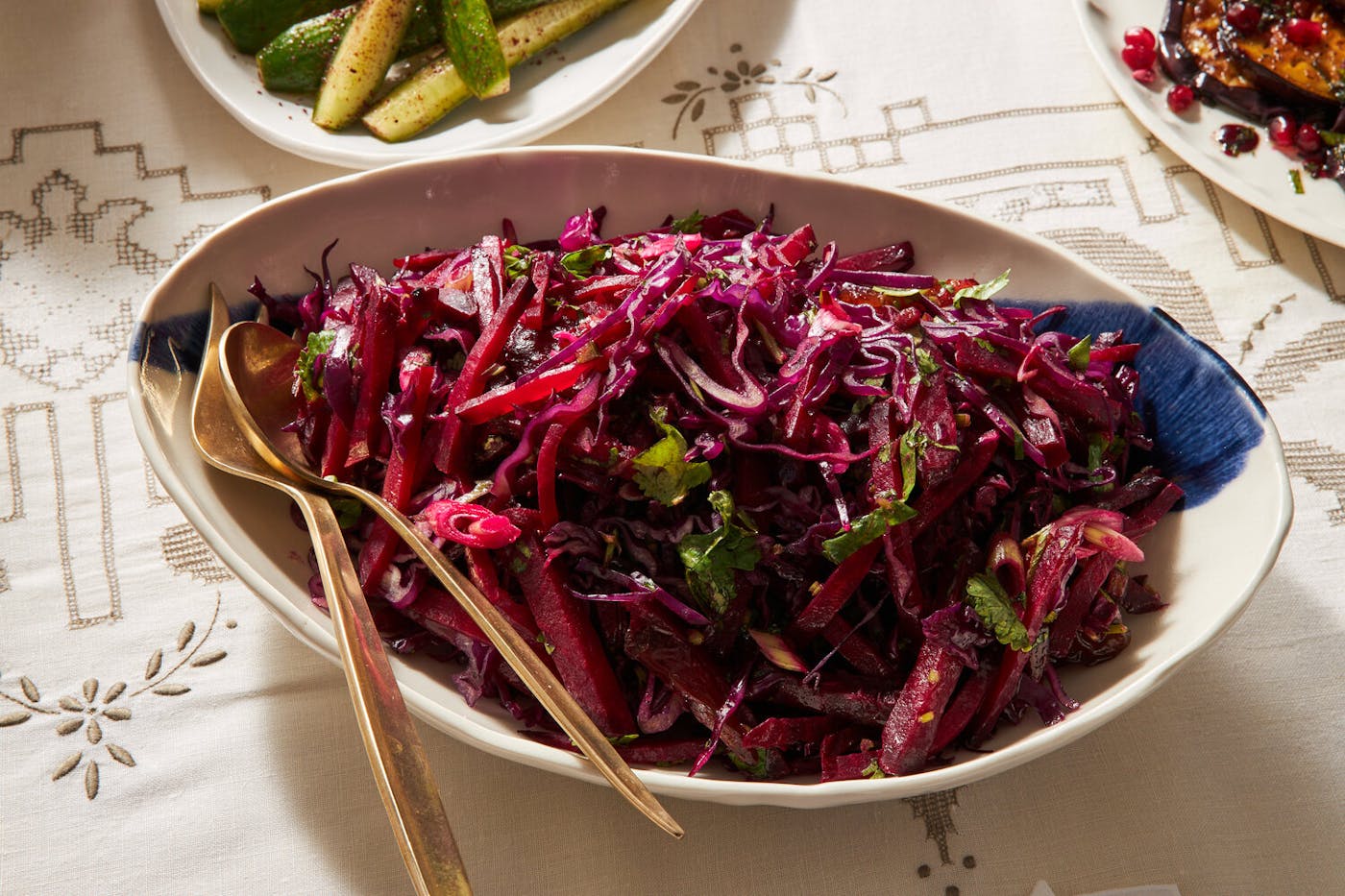 Red cabbage, date and beet salad in white bowl with gold serving utensils atop white patterned tablecloth.