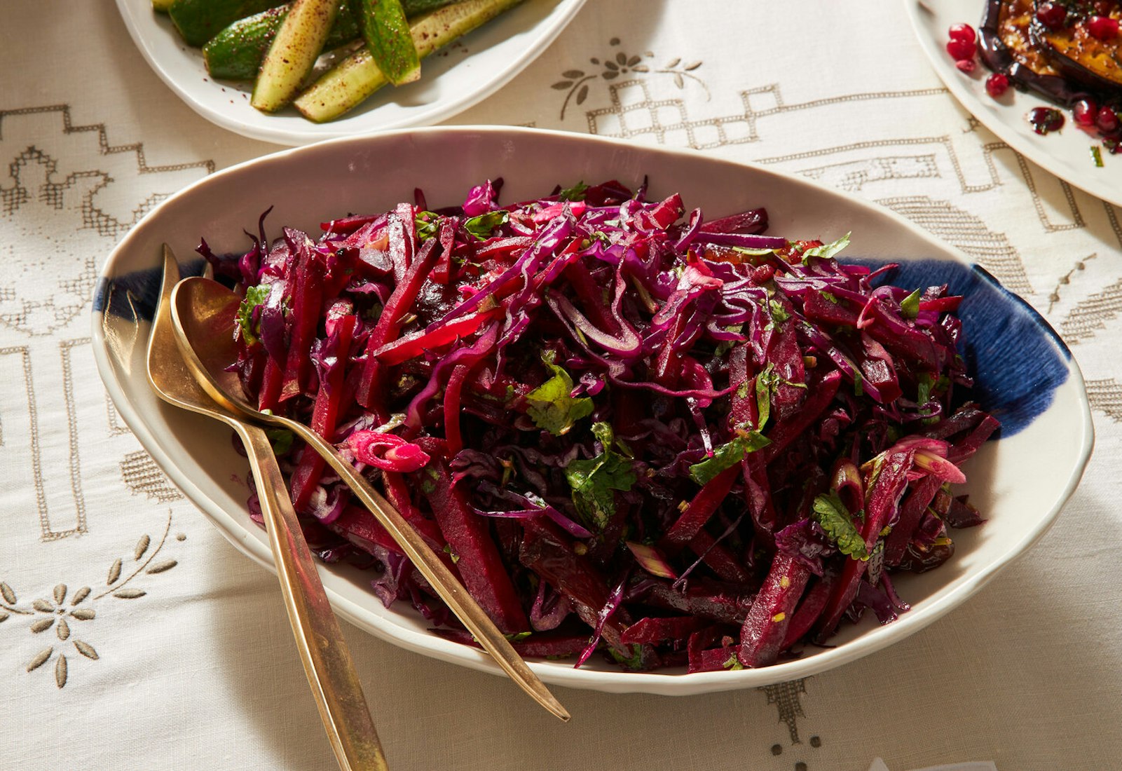 Red cabbage, date and beet salad in white bowl with gold serving utensils atop white patterned tablecloth.