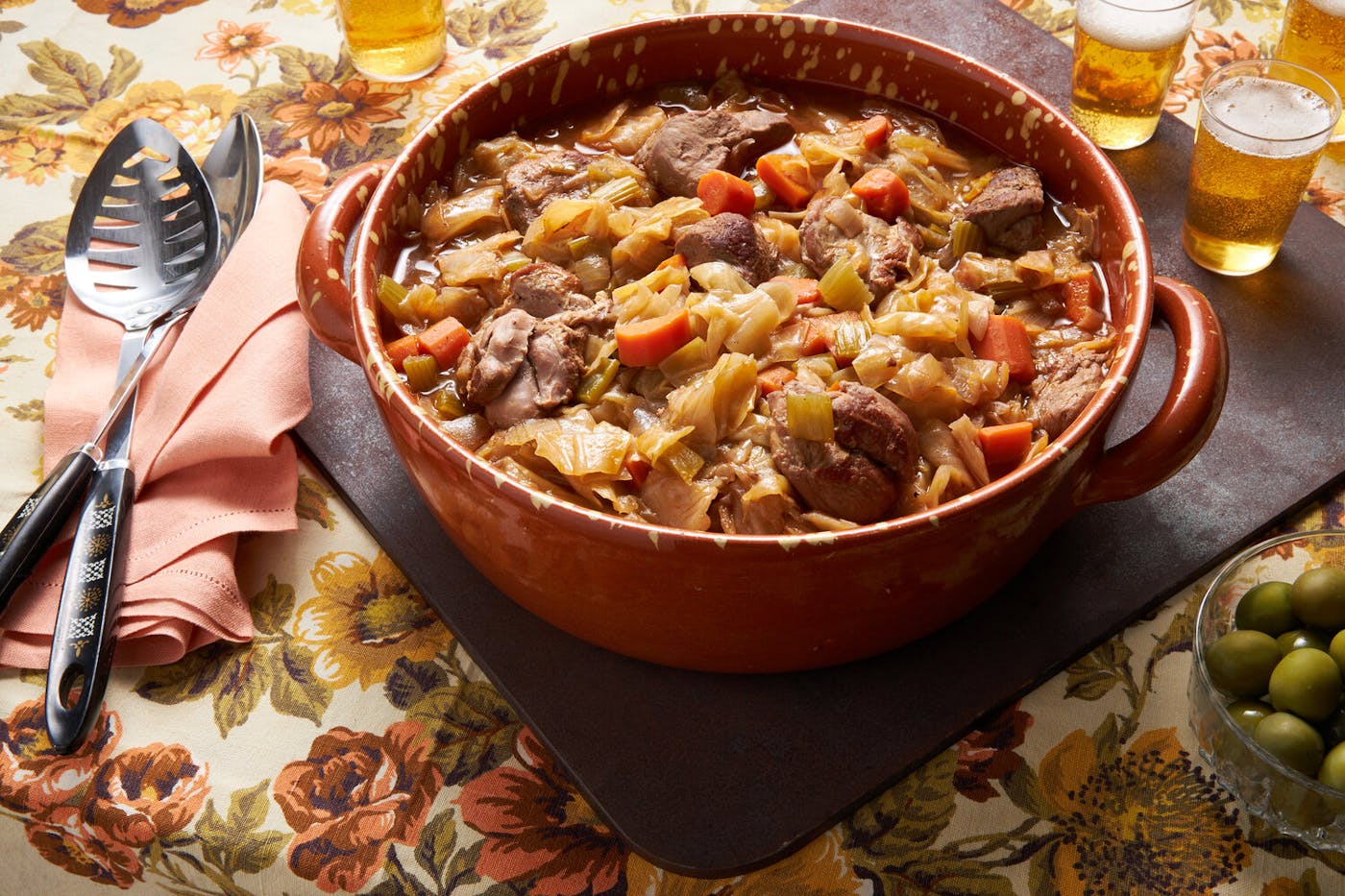 Goose cassoeula in red dutch oven, serving spoons, beer and olives atop floral tablecloth.