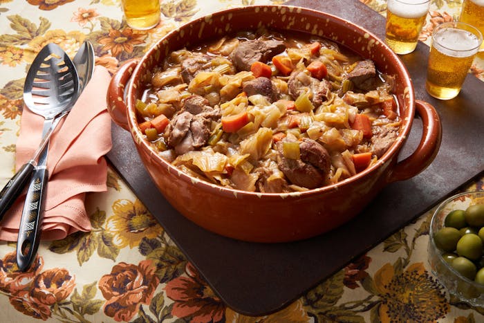 Goose cassoeula in red dutch oven, serving spoons, beer and olives atop floral tablecloth.