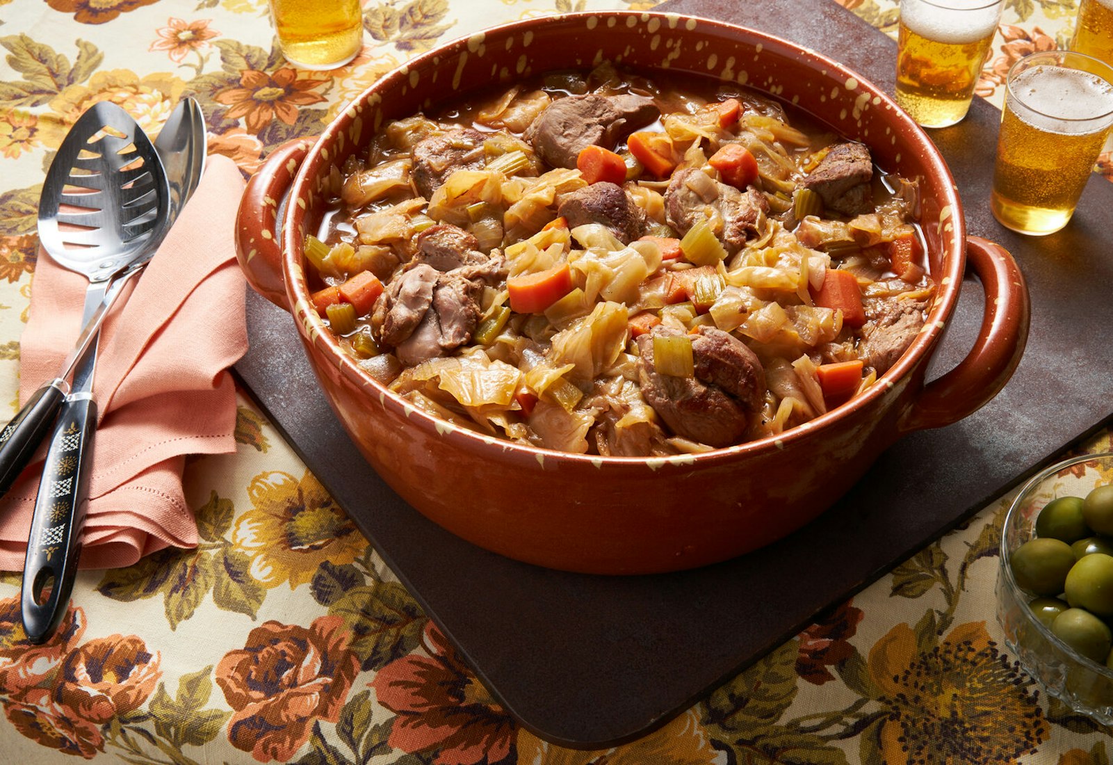 Goose cassoeula in red dutch oven, serving spoons, beer and olives atop floral tablecloth.