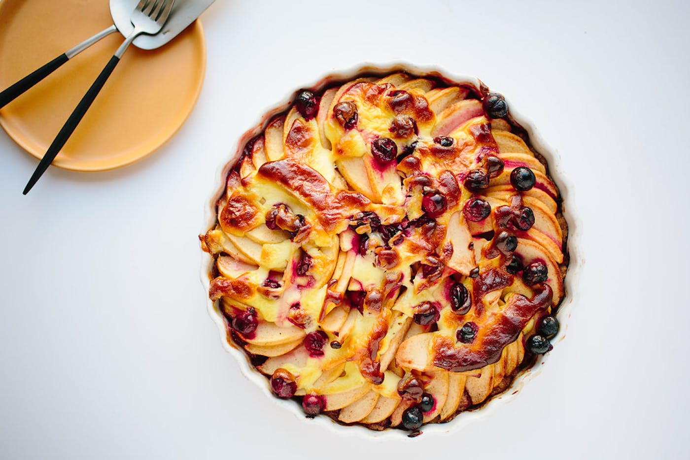 Golden apple Kuchen in scalloped cake dish alongside yellow plate with serving utensils atop white surface. 