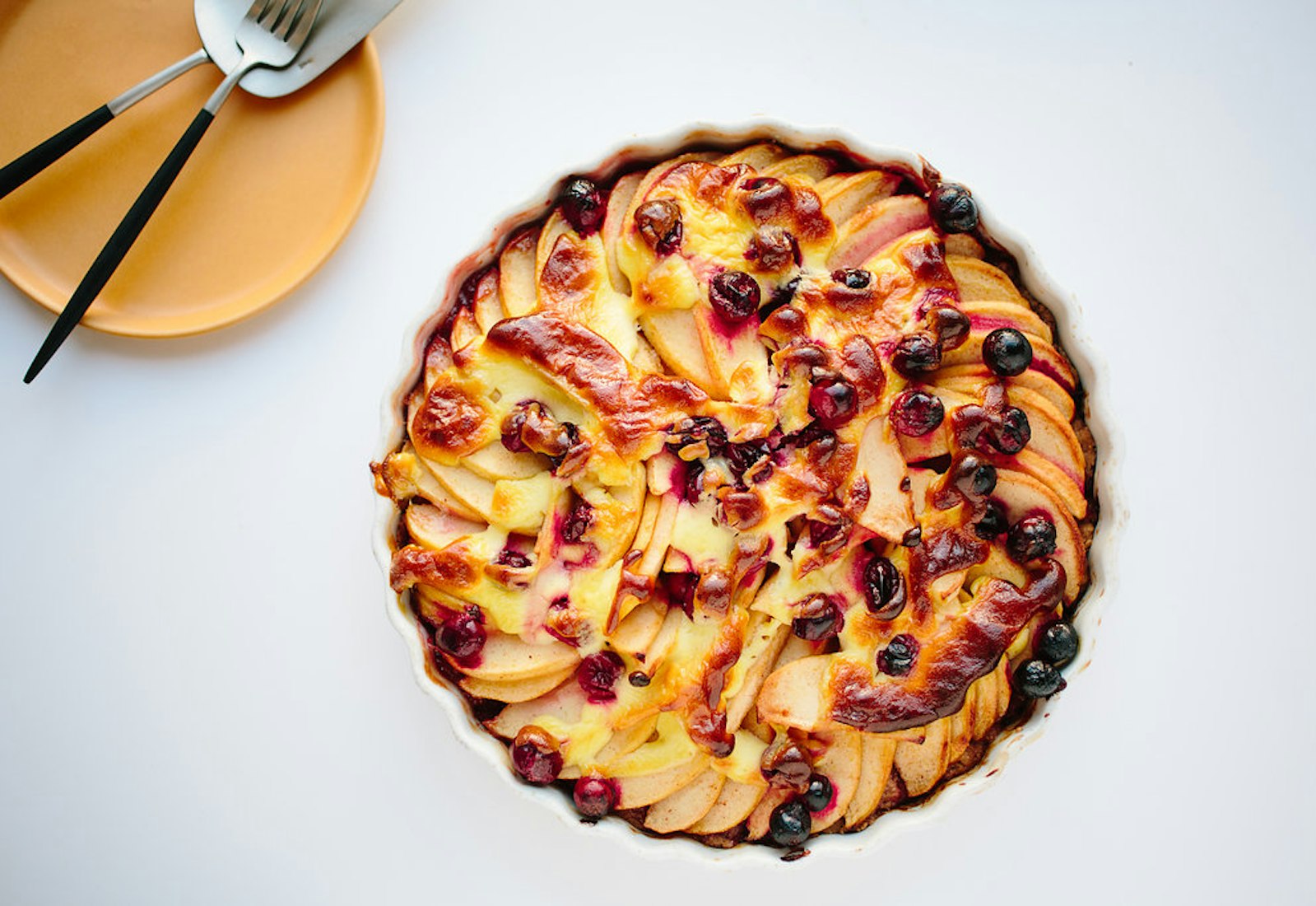 Golden apple Kuchen in scalloped cake dish alongside yellow plate with serving utensils atop white surface.