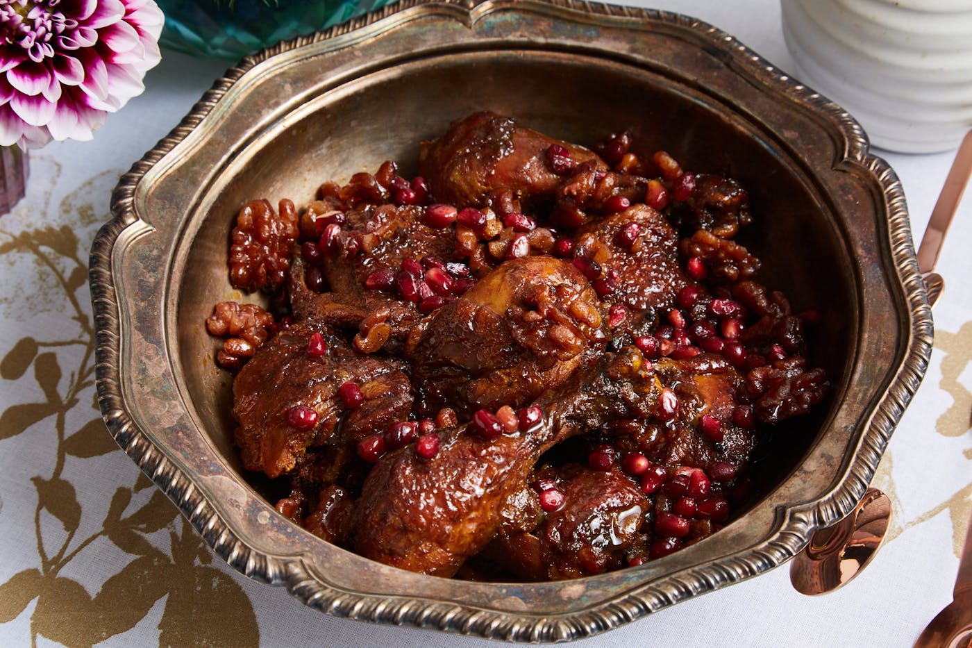 Fesenjan with fresh pomegranate kernels in bronze bowl alongside fresh flowers atop gold printed tablecloth.
