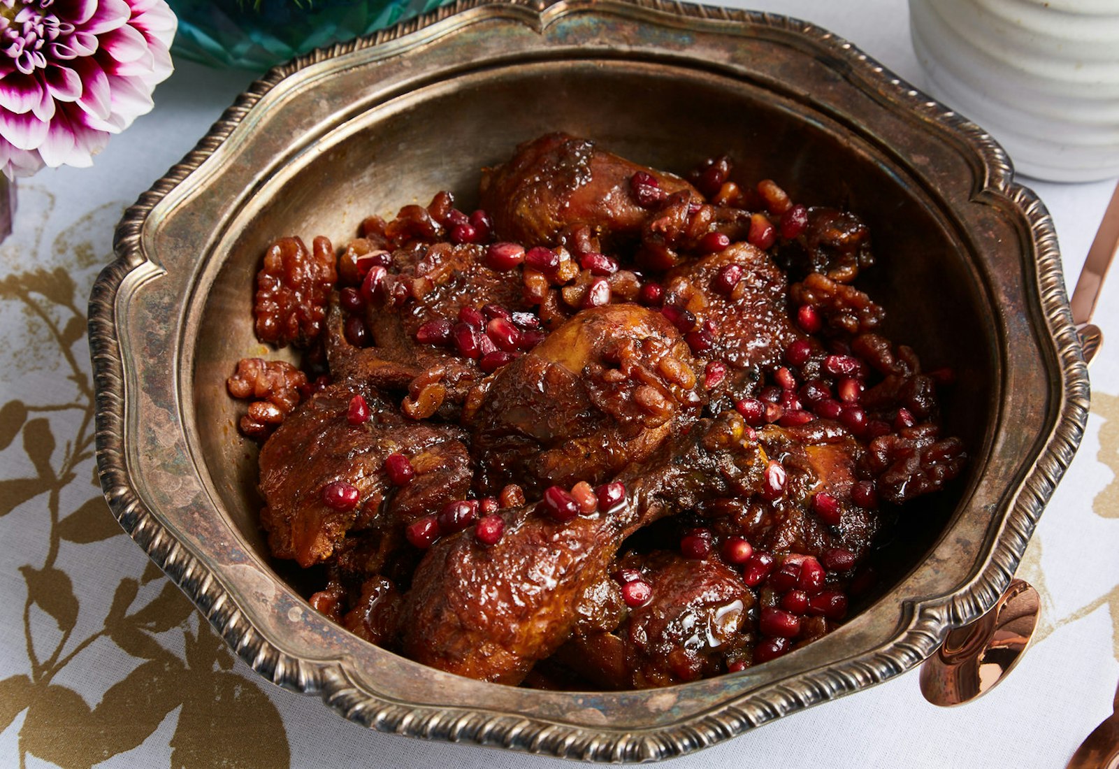 Fesenjan with fresh pomegranate kernels in bronze bowl alongside fresh flowers atop gold printed tablecloth.