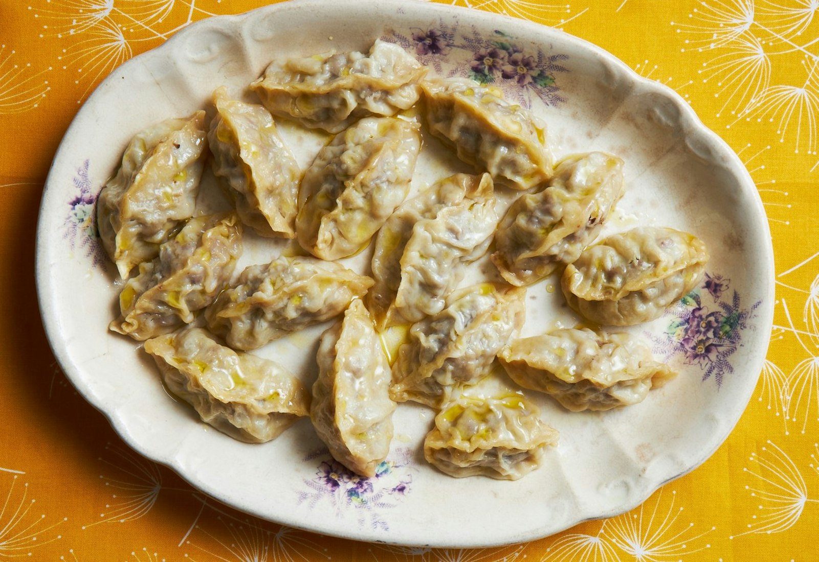 Manti drizzled with olive oil on white plate atop bright orange tablecloth.