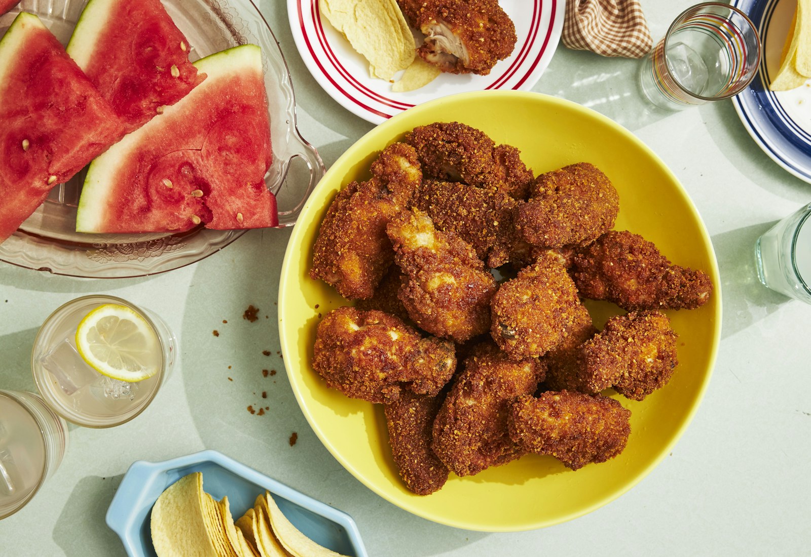 Fried chicken wings in yellow bowl next to sliced watermelon and potato chips.