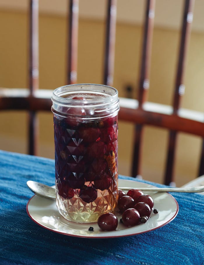 Pickled grapes with cardamom in pickling jar on white dish atop blue tablecloth.