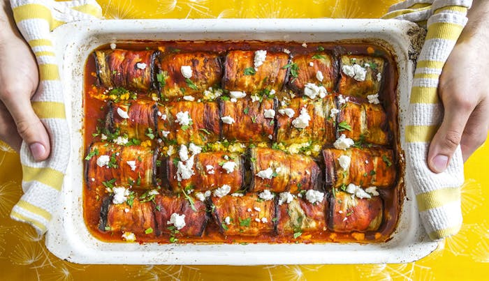 Chef holding pan of eggplant dolmas garnished with feta and chopped parsley.