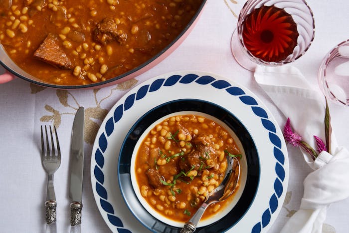Pot of yemenite stew with single serving garnished with cilantro garnish on a blue and white plate, glass of red wine and pink flowers.
