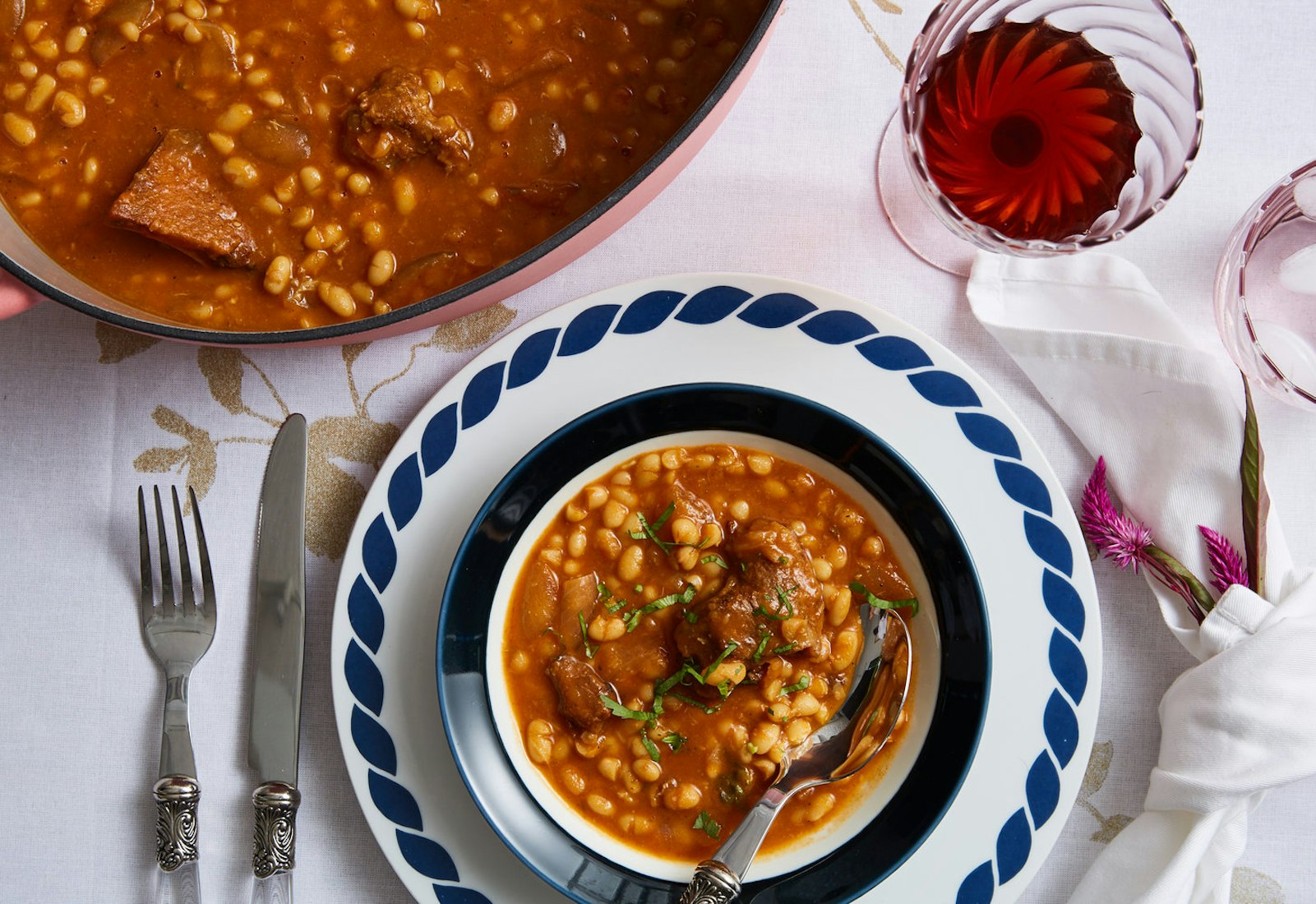 Pot of yemenite stew with single serving garnished with cilantro garnish on a blue and white plate, glass of red wine and pink flowers.
