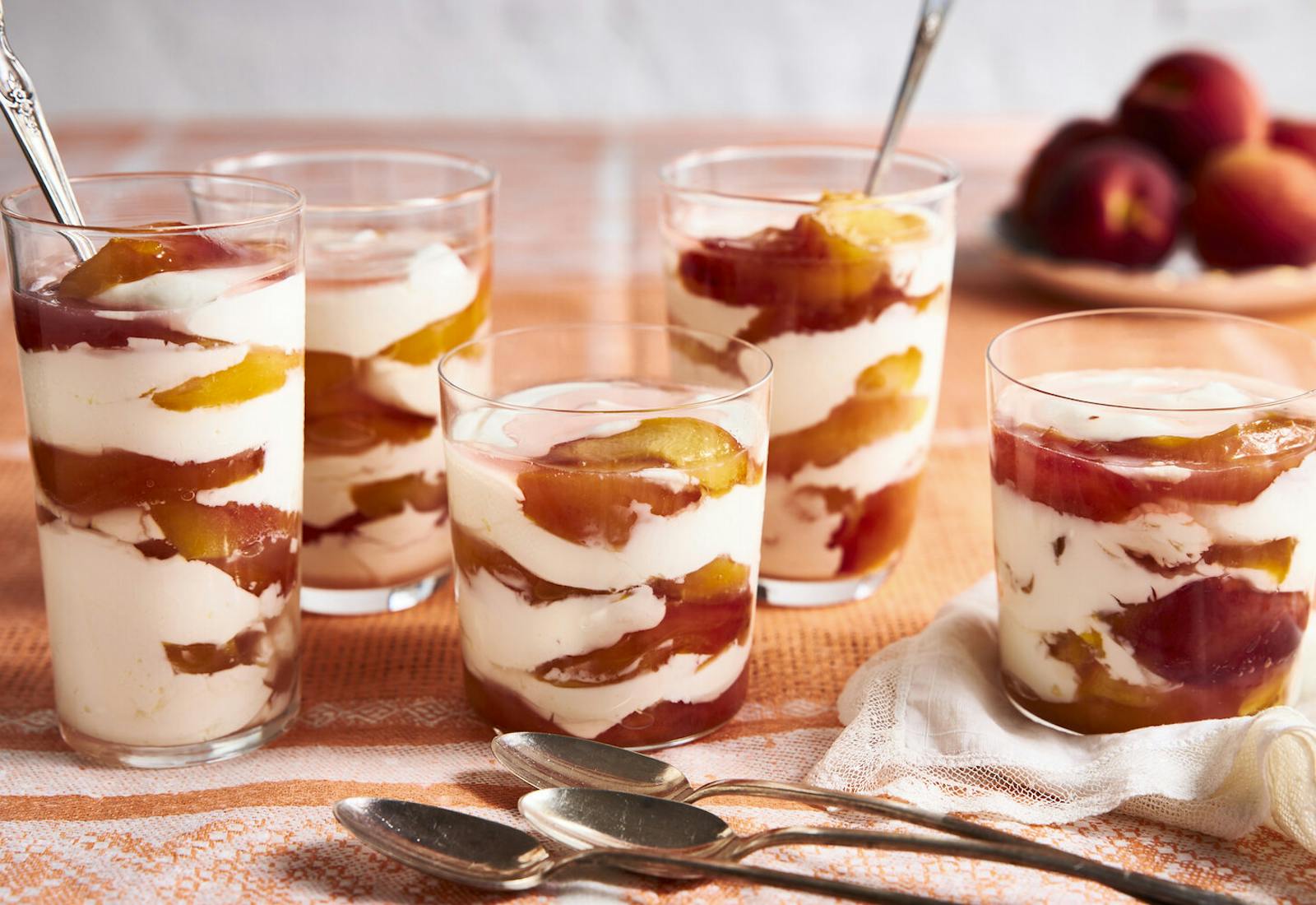 Parfait cups with layers of peaches and sour cream with bowl of fresh peaches in the background atop orange tablecloth.
