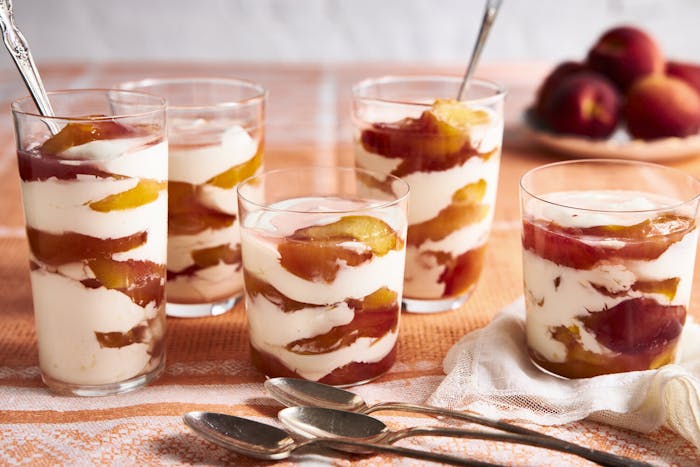 Parfait cups with layers of peaches and sour cream with bowl of fresh peaches in the background atop orange tablecloth.