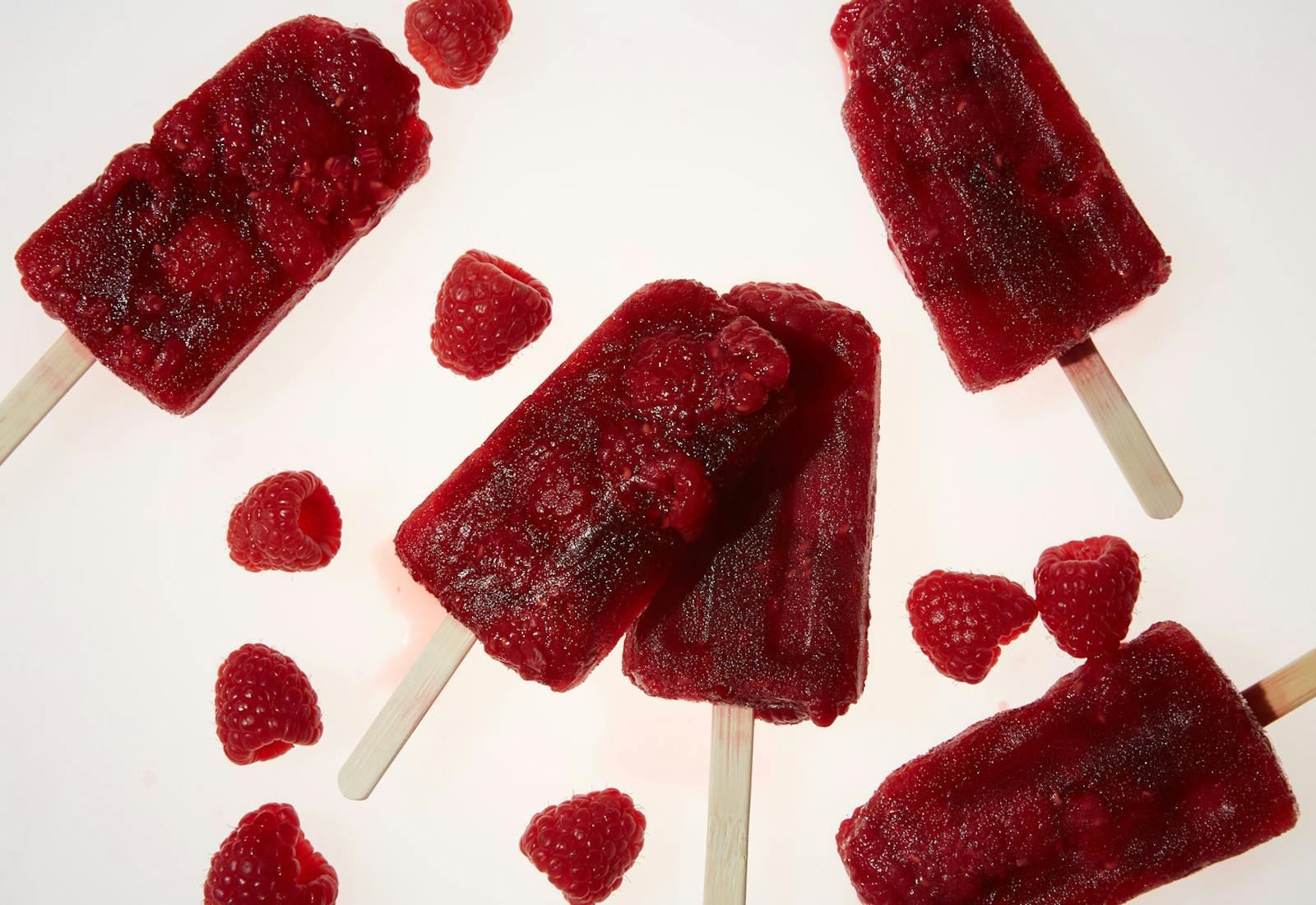 Raspberry ice pops with scattered fresh raspberries on white surface.