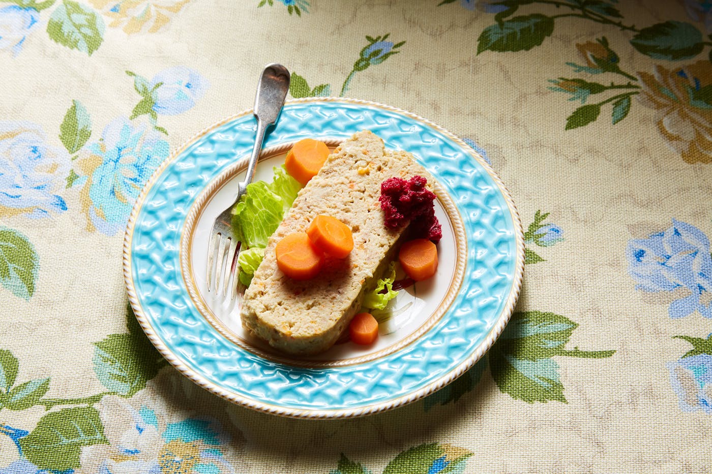 Slice of gefilte fish with carrots, beet horseradish and lettuce on blue plate atop floral tablecloth.