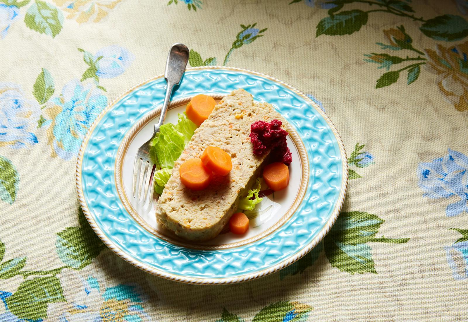 Slice of gefilte fish with carrots, beet horseradish and lettuce on blue plate atop floral tablecloth.