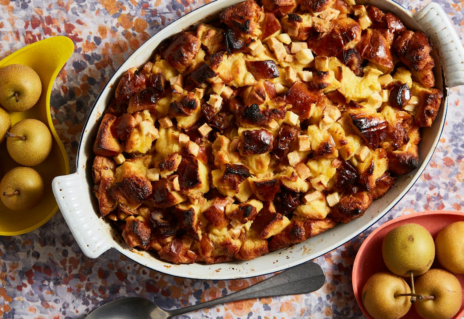 Apple and date challah bread pudding in white casserole dish alongside plates of fresh apples, atop floral tablecloth.