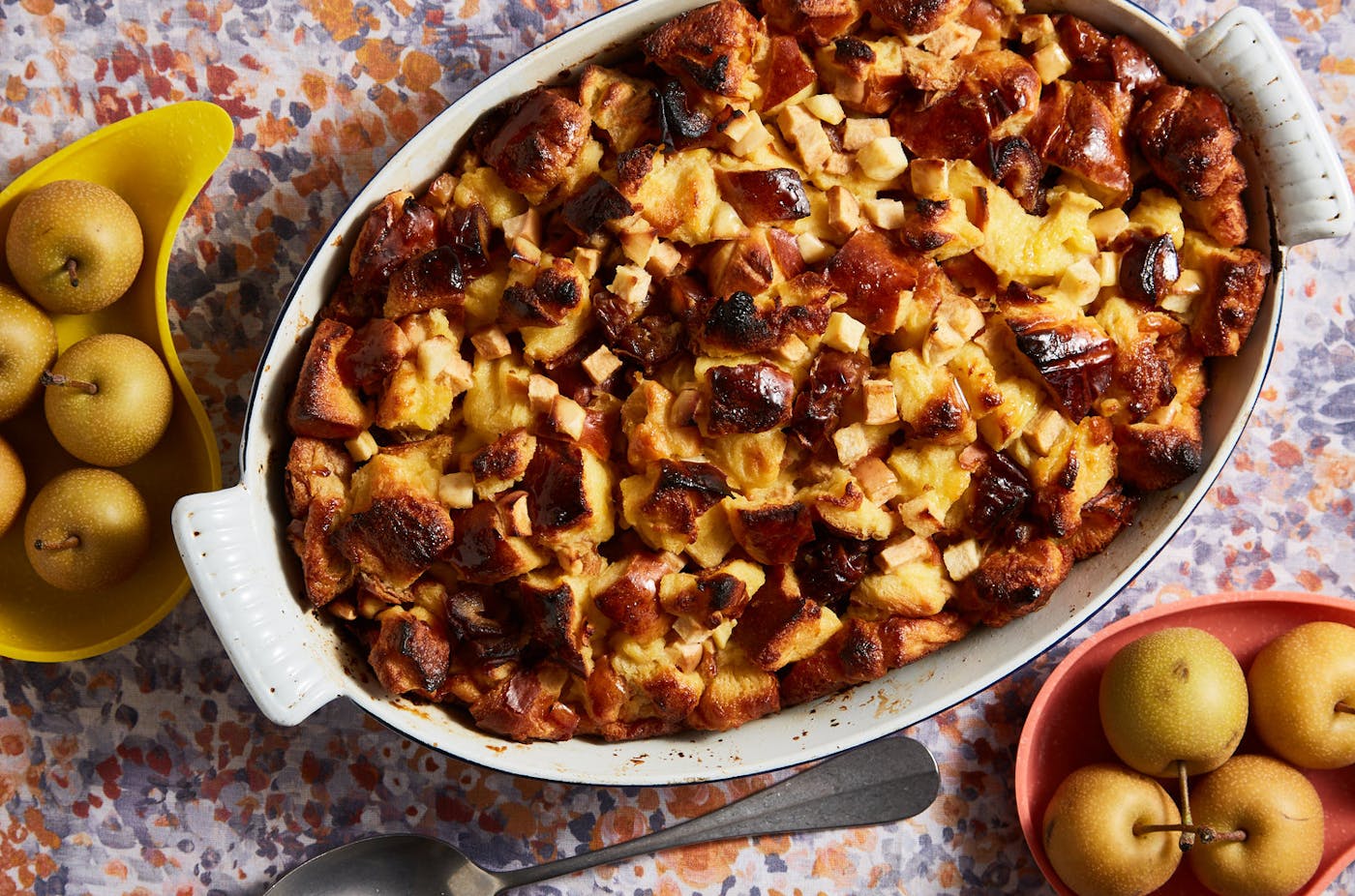 Apple and date challah bread pudding in white casserole dish alongside plates of fresh apples, atop floral tablecloth.
