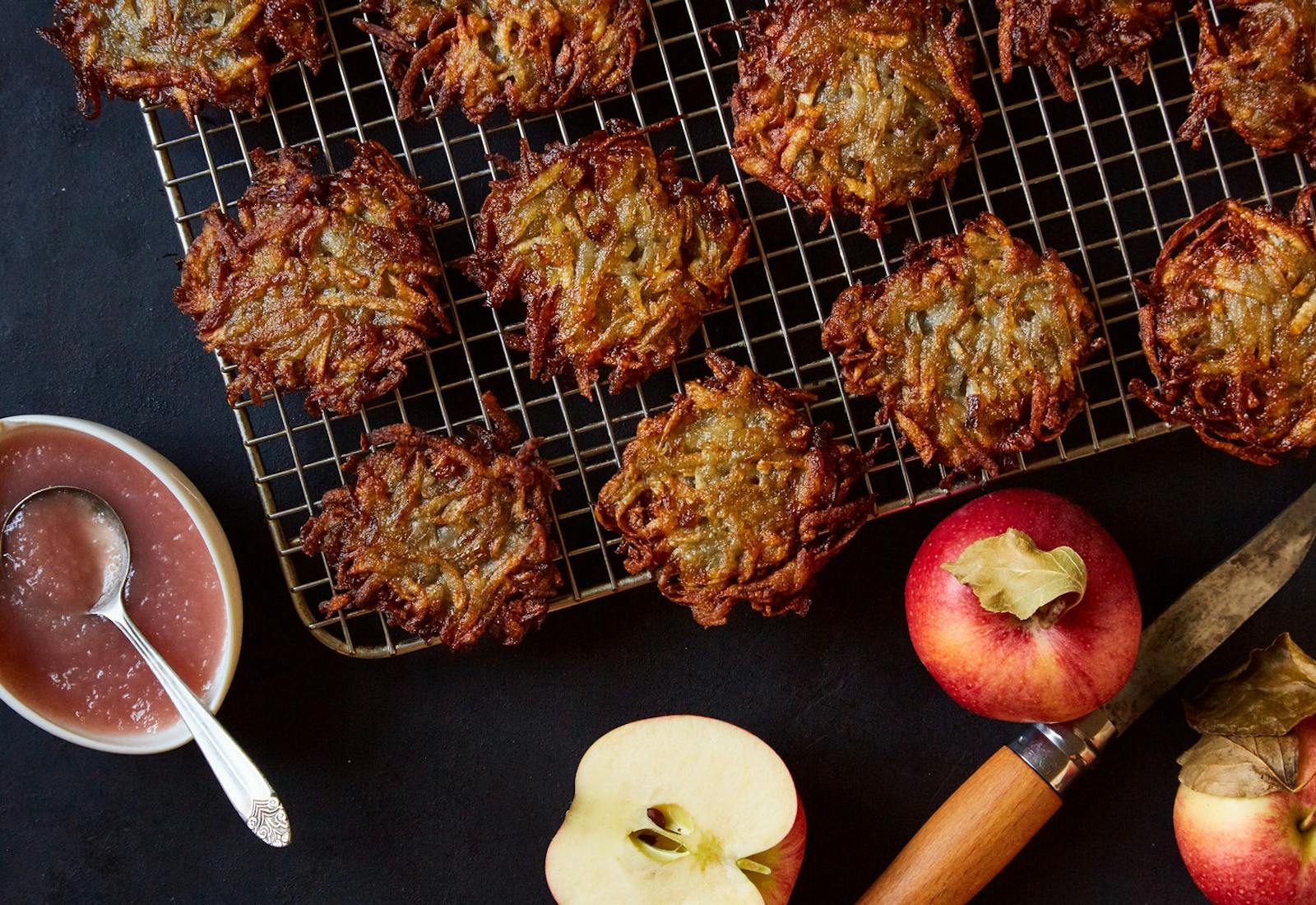 Latkes on wire baking rack alongside apple sauce and fresh apples atop black surface.