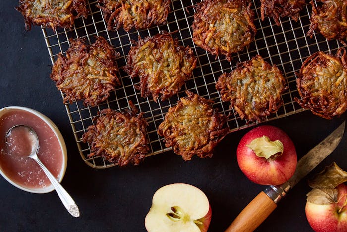 Latkes on wire baking rack alongside apple sauce and fresh apples atop black surface.