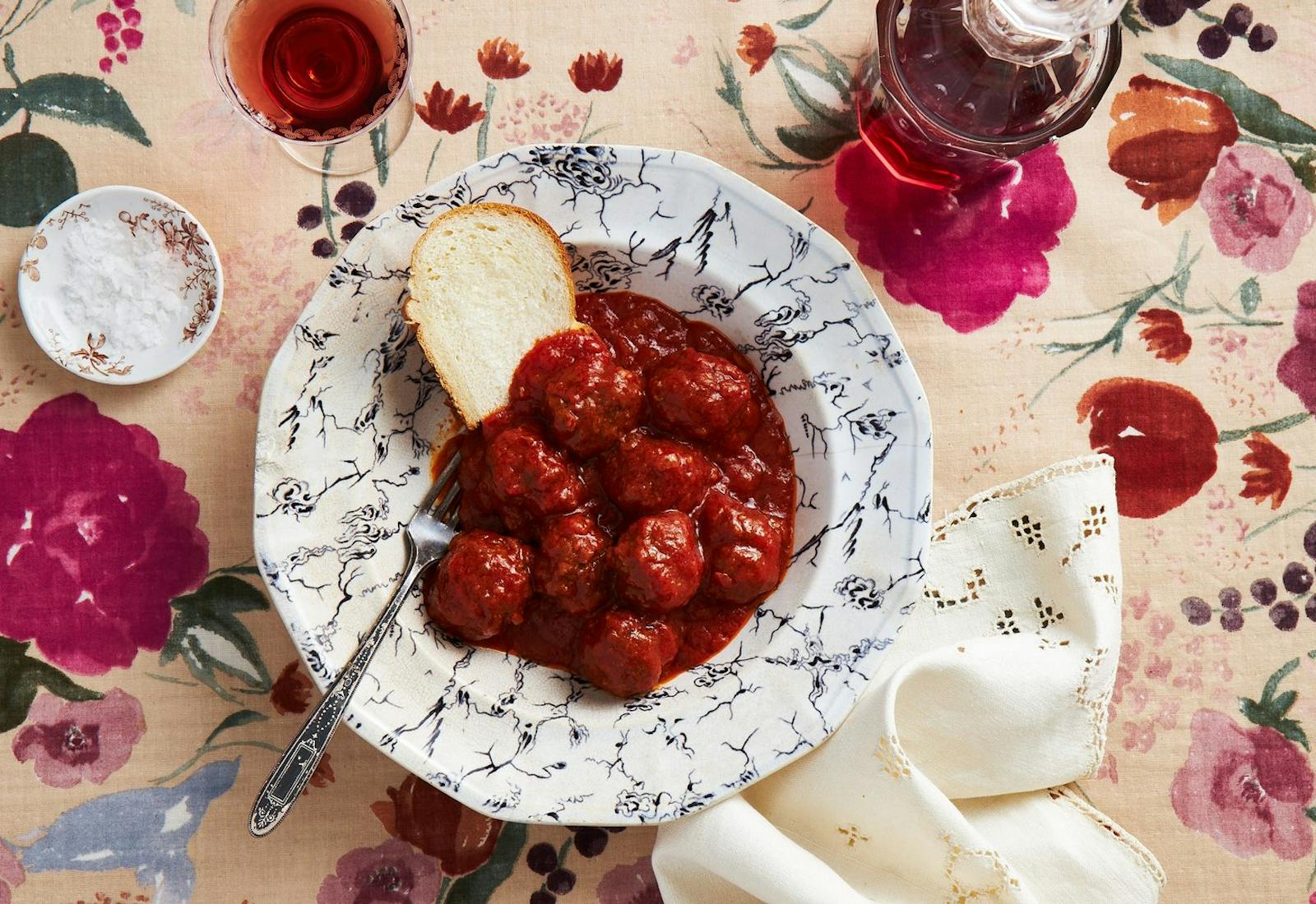 Meatballs and tomato sauce with challah and red wine atop a floral tablecloth.