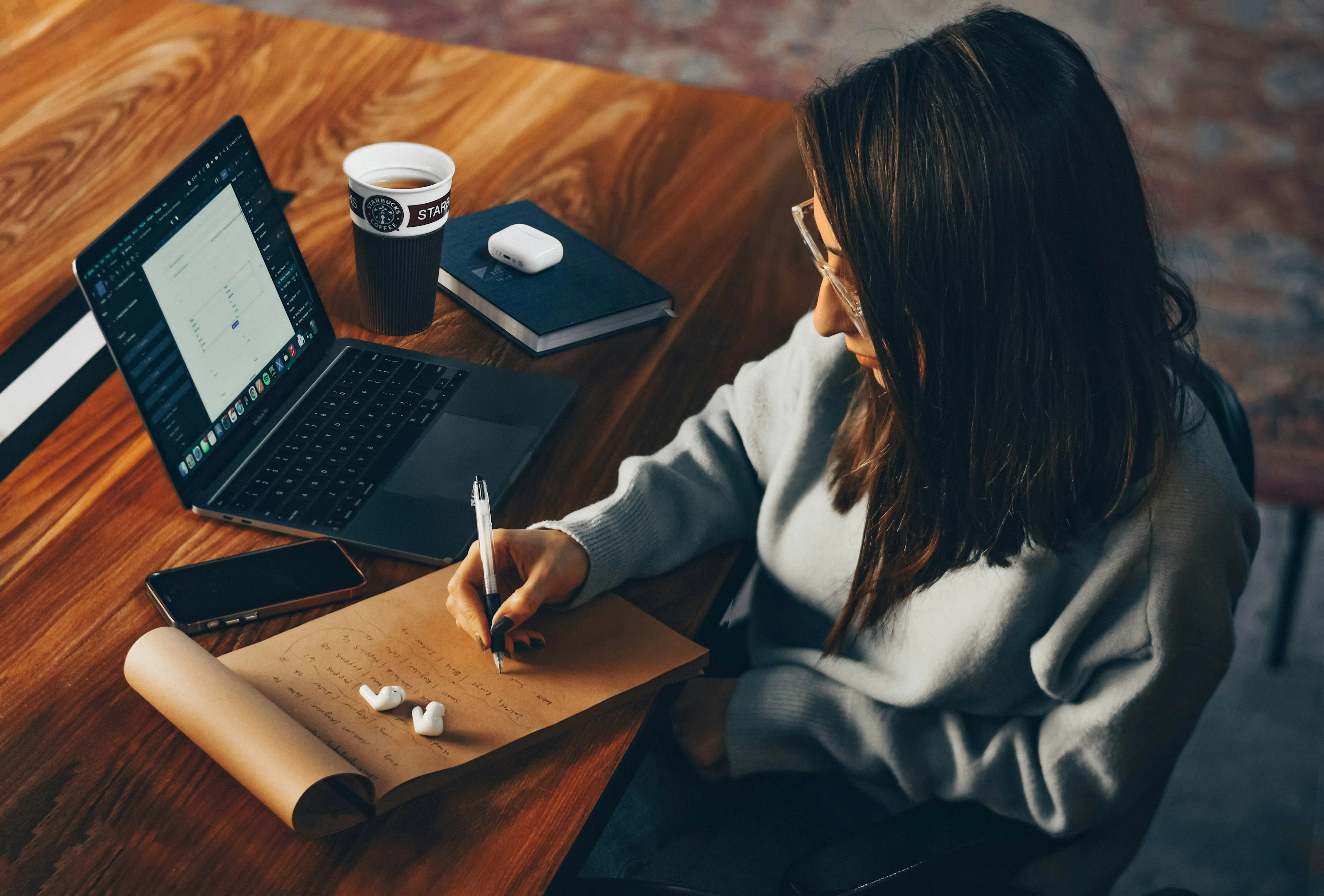 side profile of a woman wearing glasses and shoulder length brown hair sitting at a desk in front of her laptop with a cup of coffee with her phone a notebook and airpods while writing on a notepad.