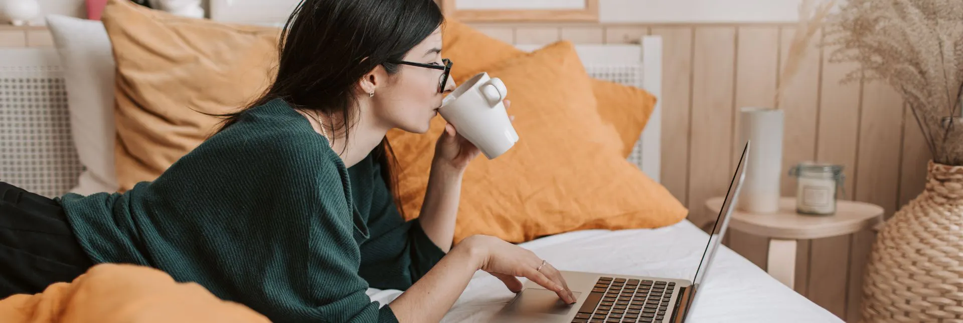 A person laying on their bed drinking coffee and using a laptop