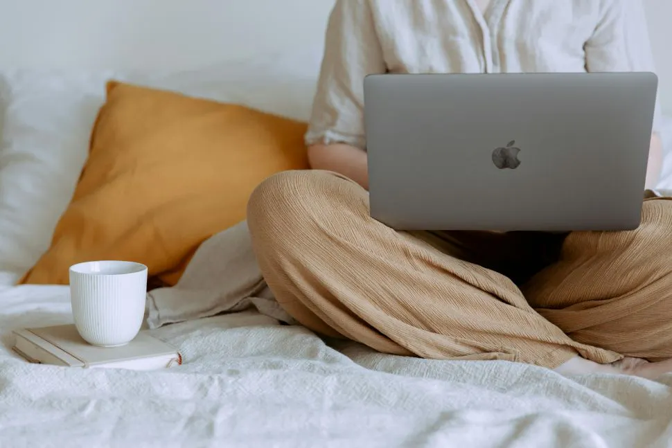 A person sitting on a bed drinking coffee and using a laptop