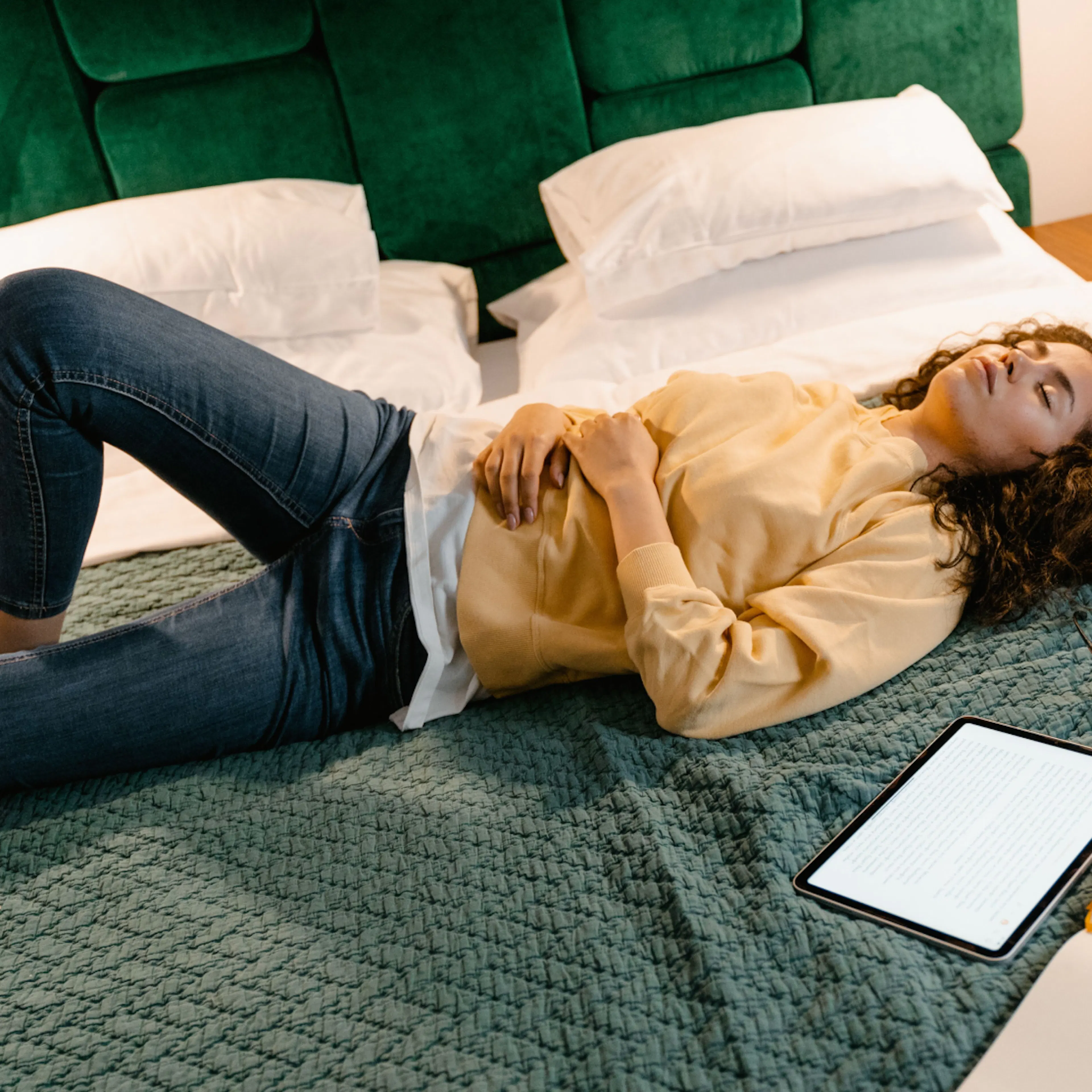 A woman lying down on top of a green comforter, holding her stomach and looking uncomfortable. She may be experience estrogen dominance symptoms like bloating or worsening PMS.