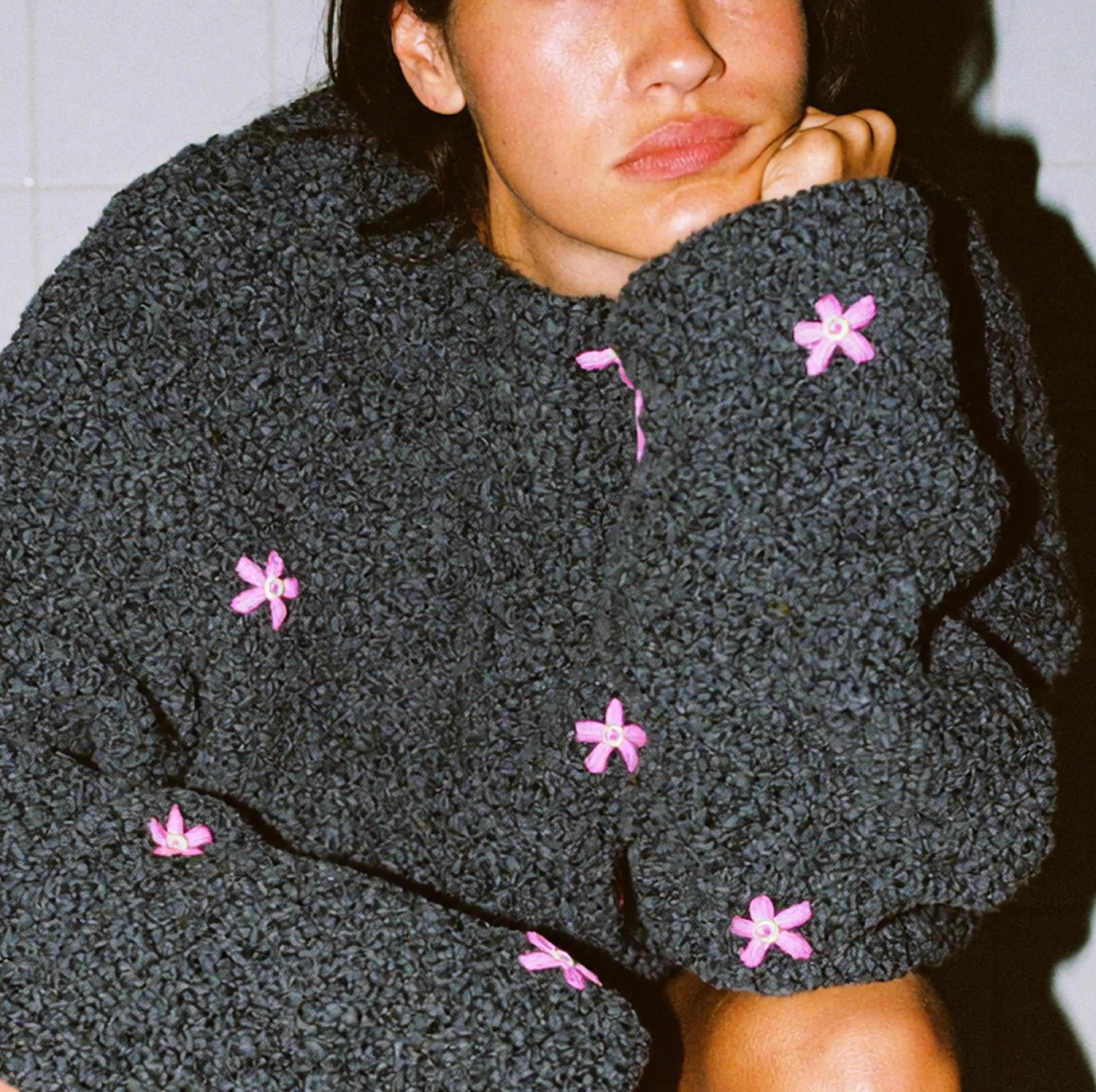 A close-up image of a woman frowning with her arm resting on her knee and her head resting in her hand. She's wearing a gray hoodie with pink flowers on it.