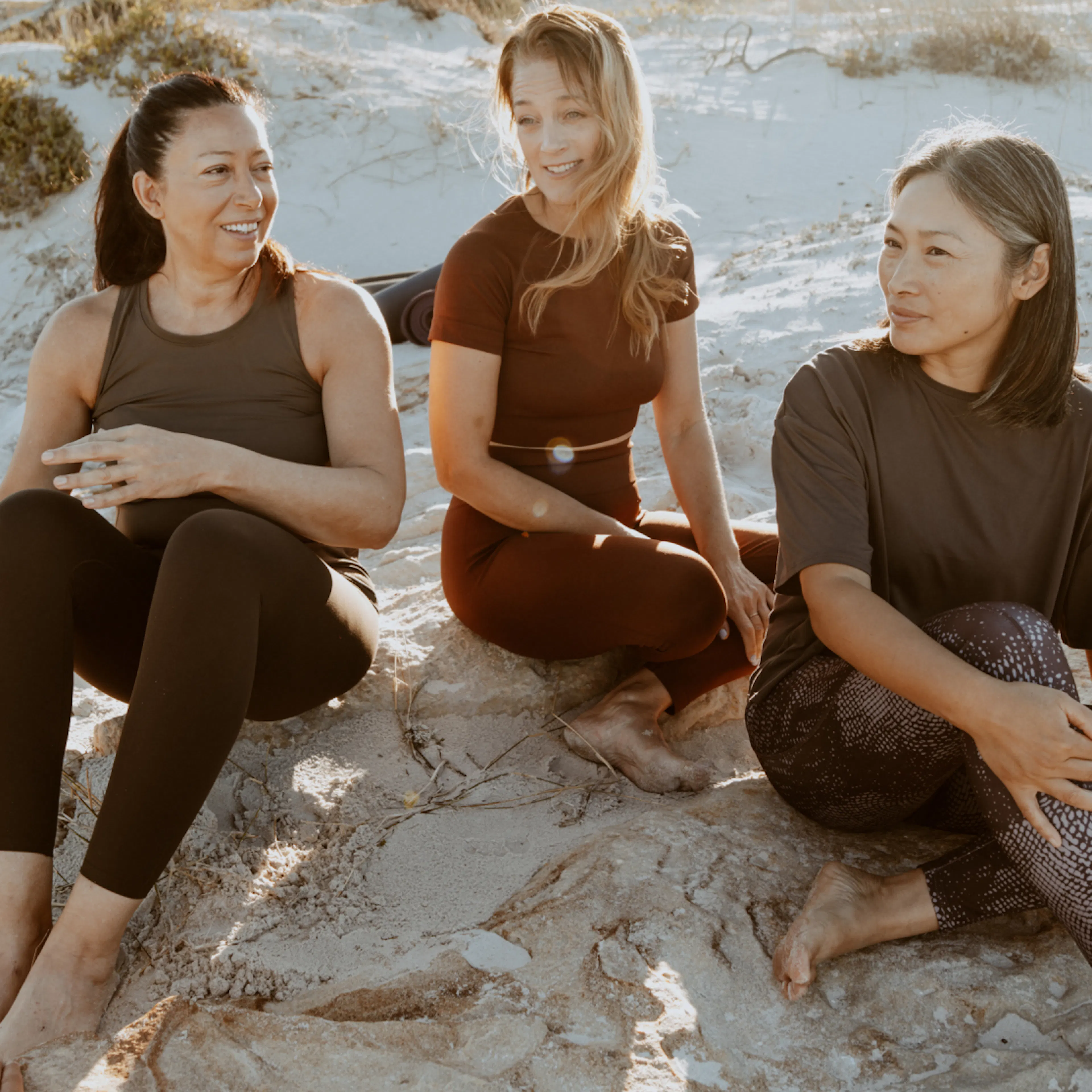 Three women in midlife, sitting on the sand together and relaxing.