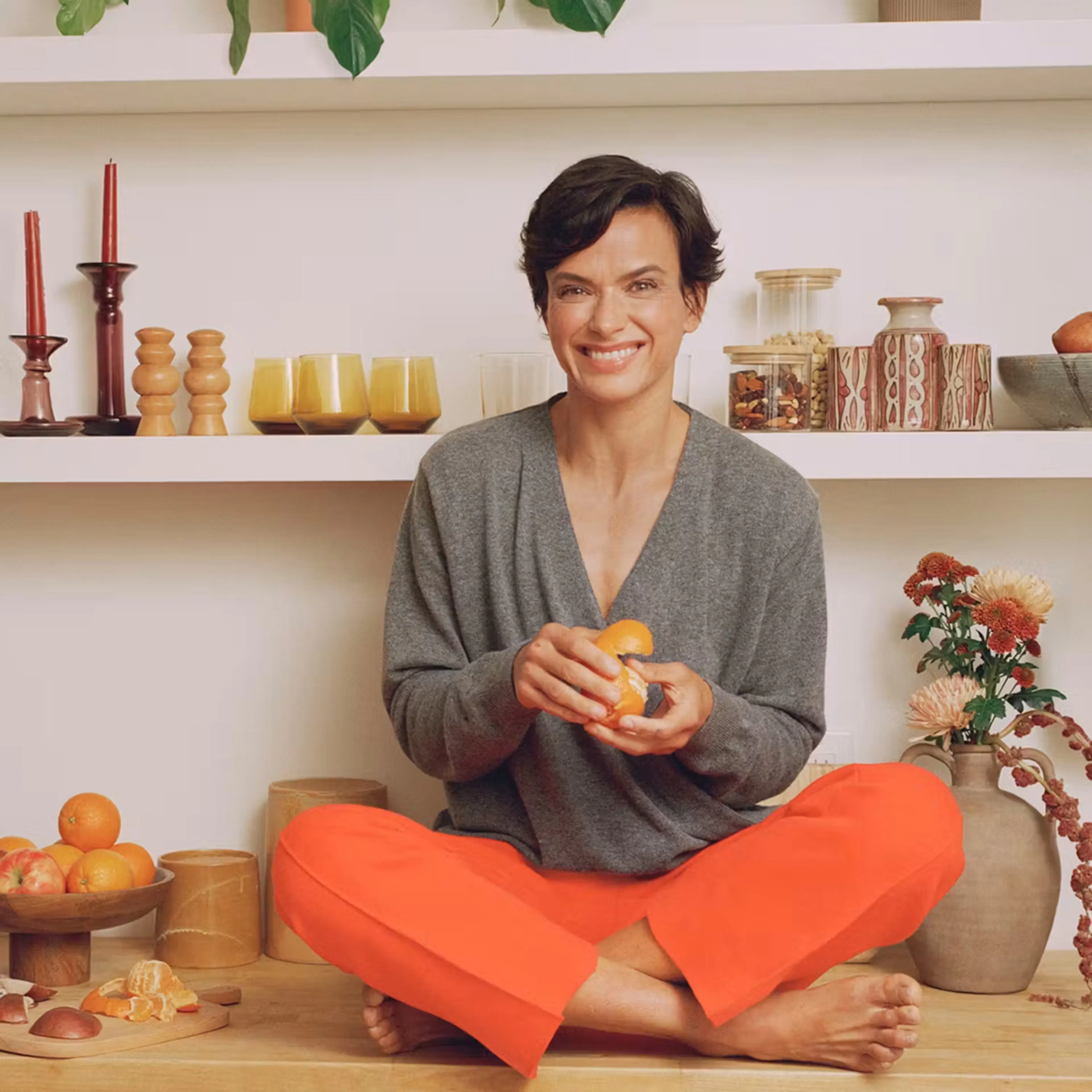 Smiling brunette woman with orange pants sitting while peeling an orange