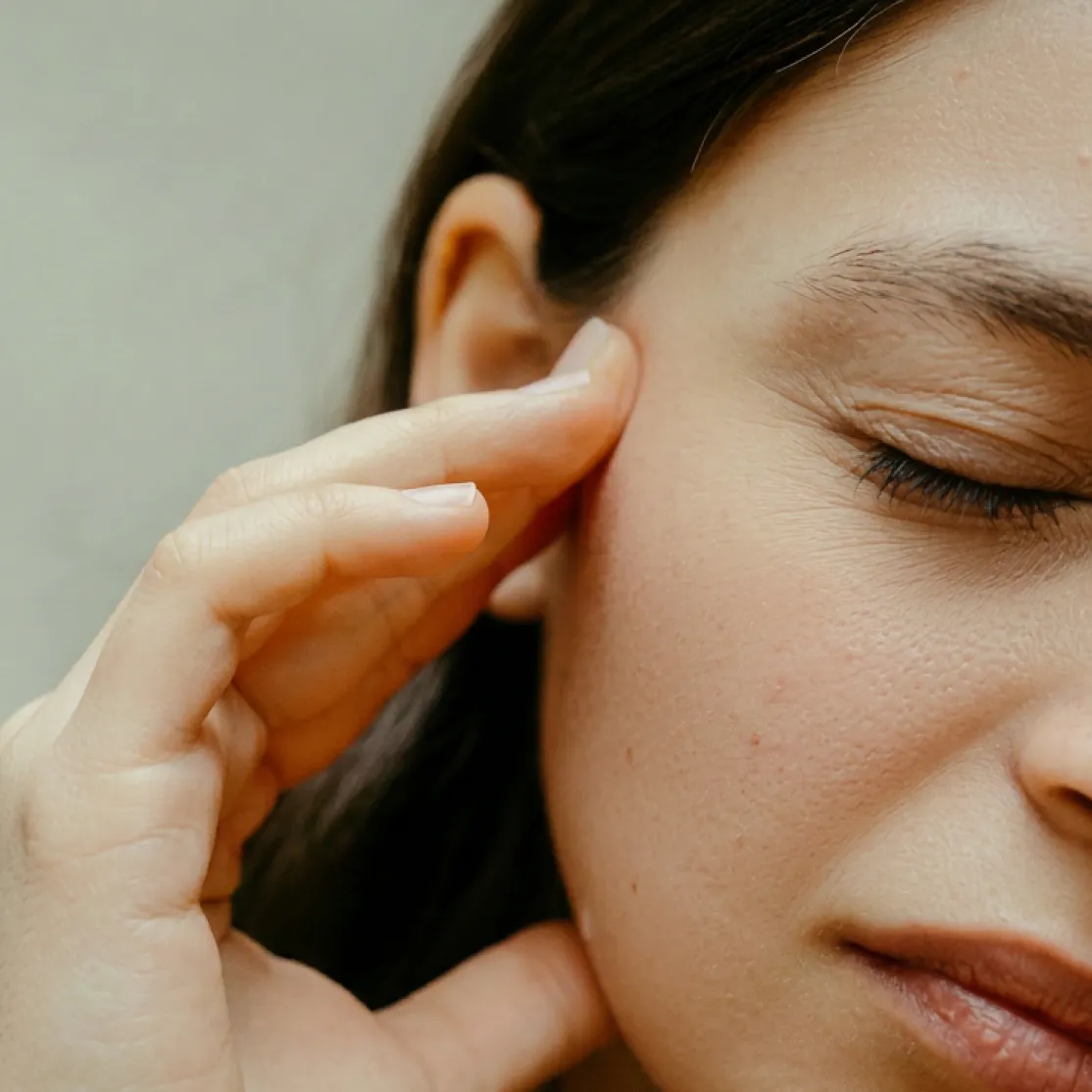 A close-up photo of a woman in menopause experiencing tinnitus. She's holding her head and closing her eyes.