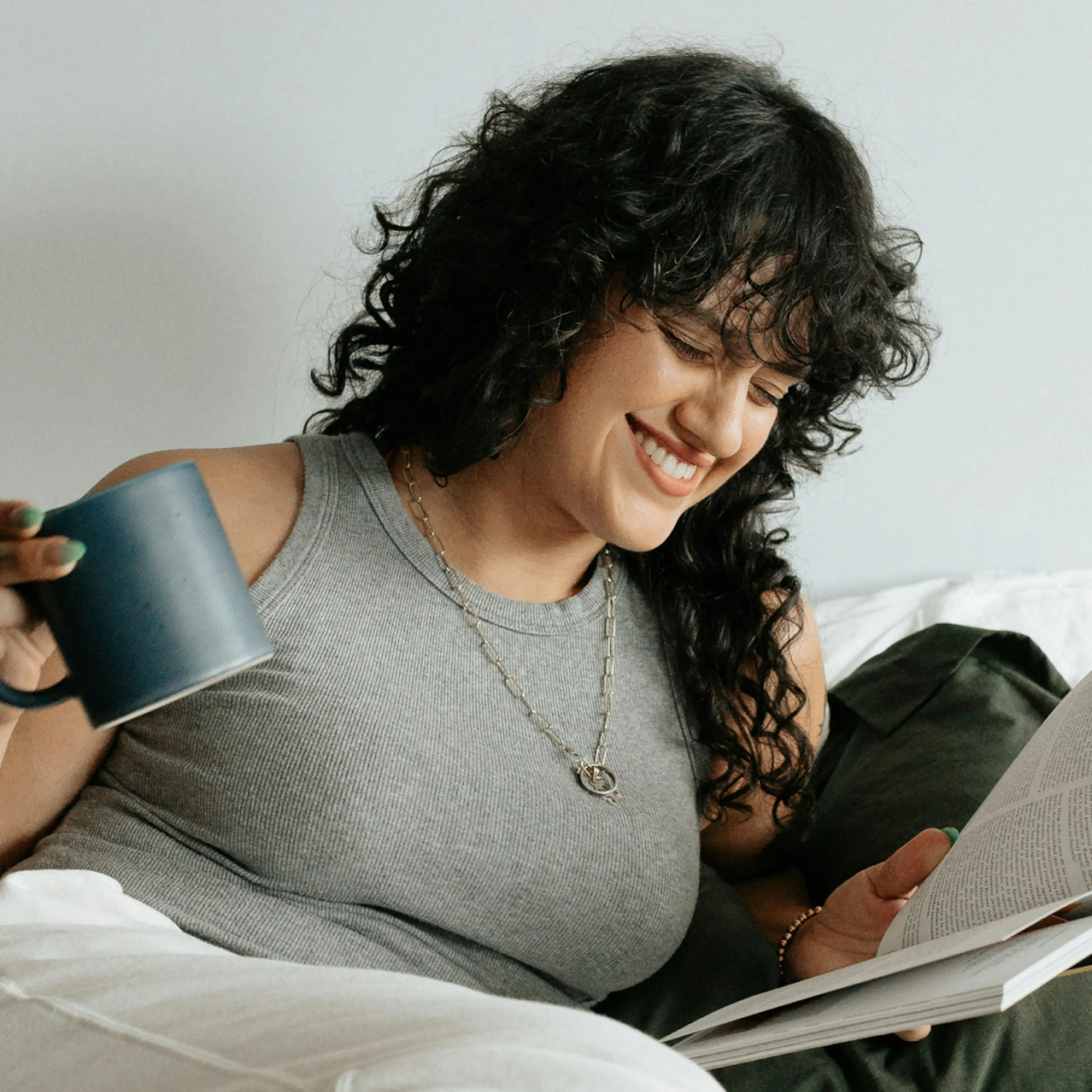A woman in perimenopause smiling, reading something, and holding a mug while relaxing inside.
