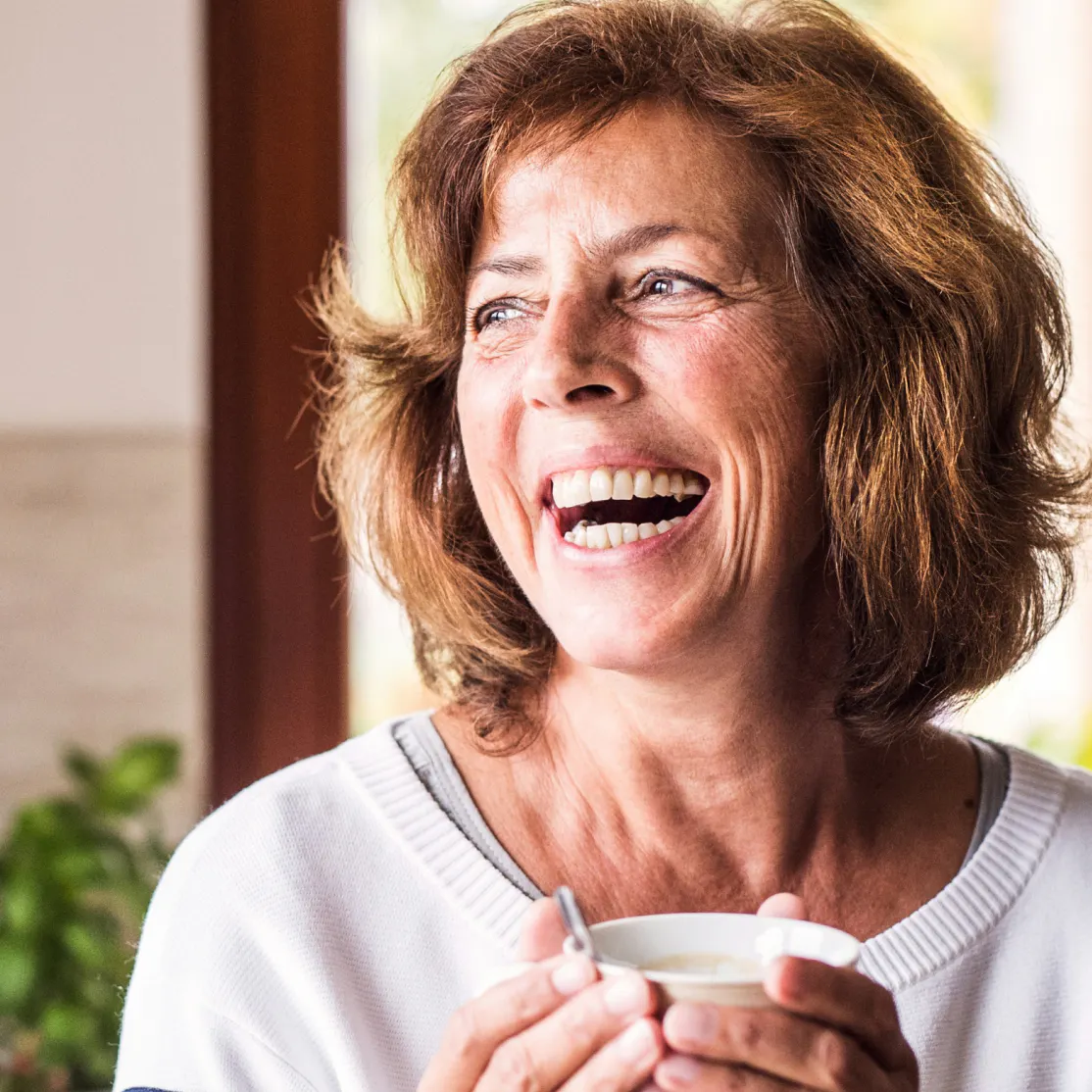 A woman in menopause smiling and laughing, turning to the right and holding a coffee mug.