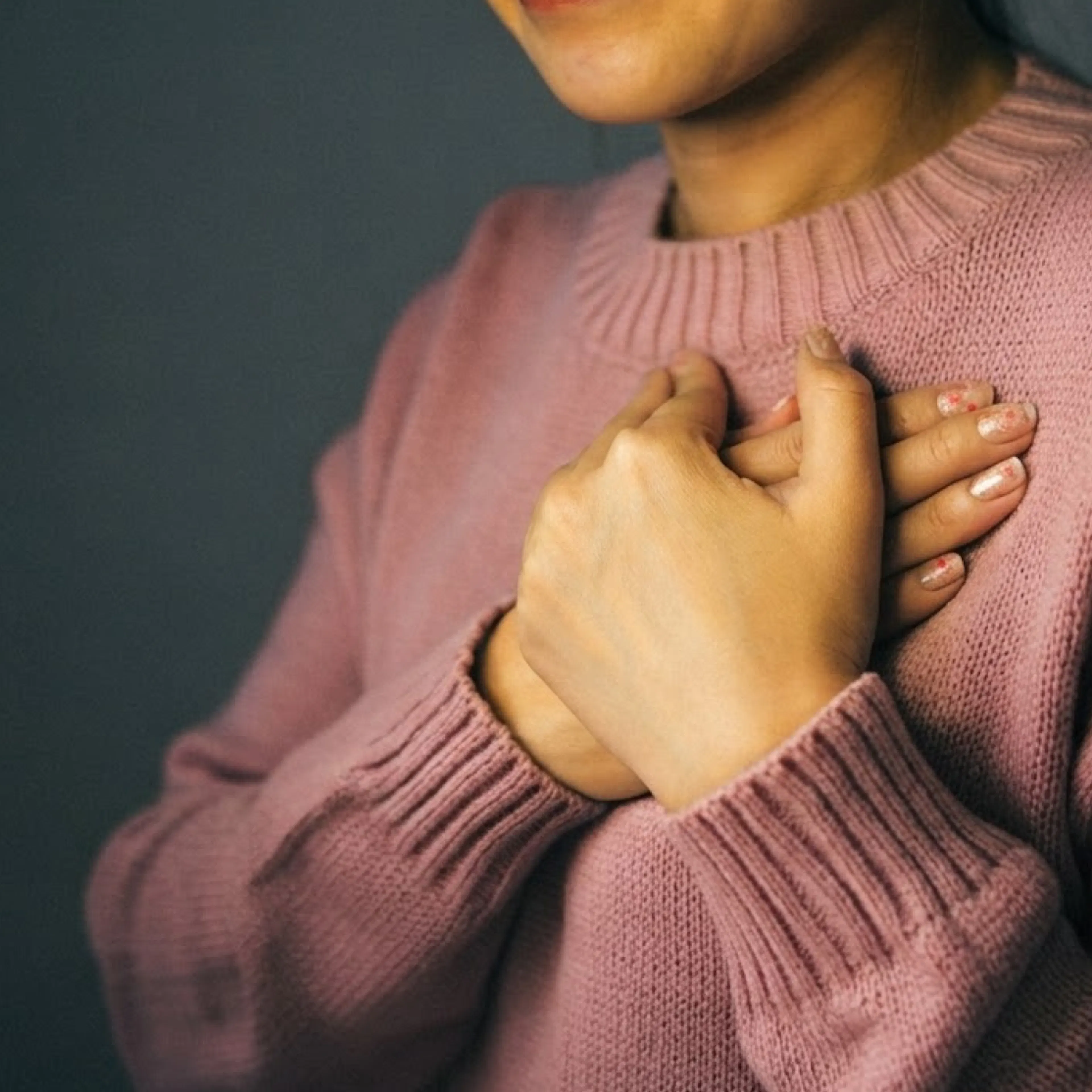 A close-up image of a woman's hands resting on her chest over her heart. She's wearing a pink sweater.
