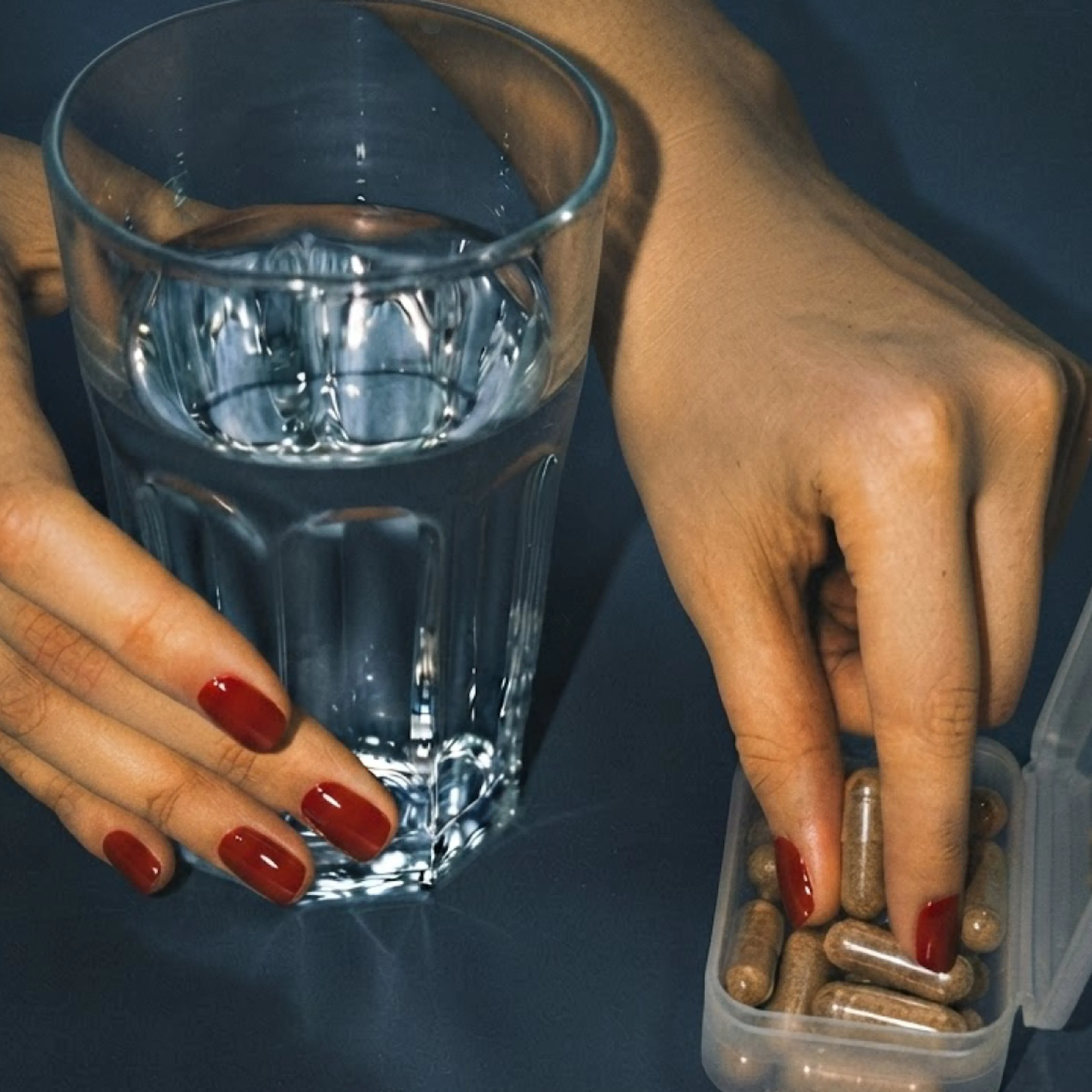 A woman's hand holding onto a glass of water and a pill box containing brown supplements.