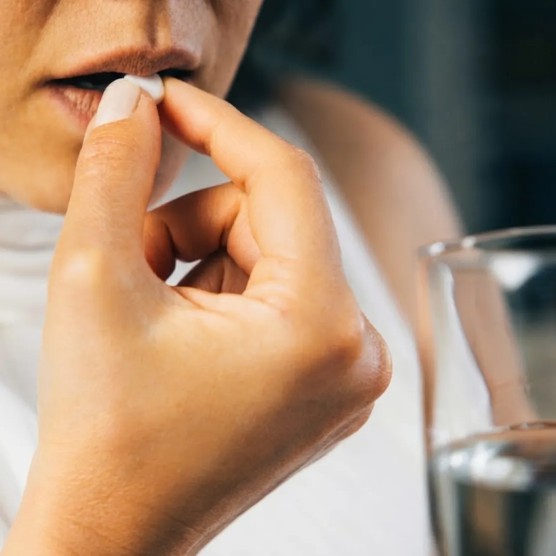 A close-up image of a woman in midlife taking a pill with a glass of water.